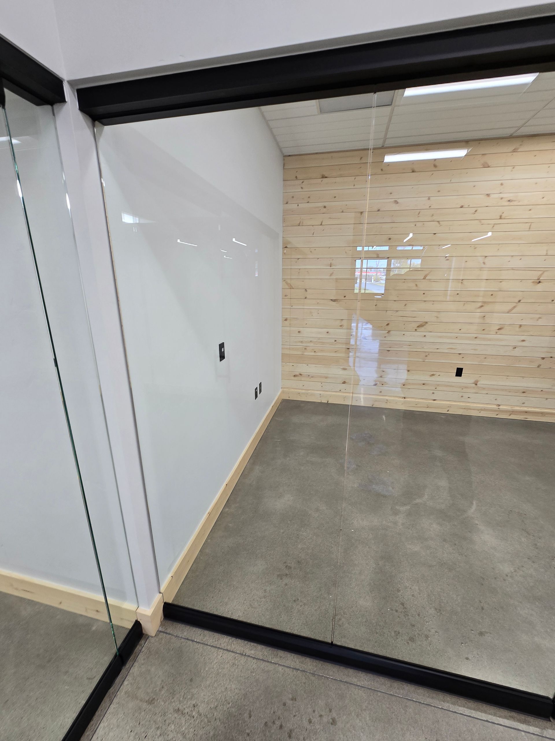 A modern office space features a glass partition, polished concrete floors, white walls, and a light-wood panel accent wall.
