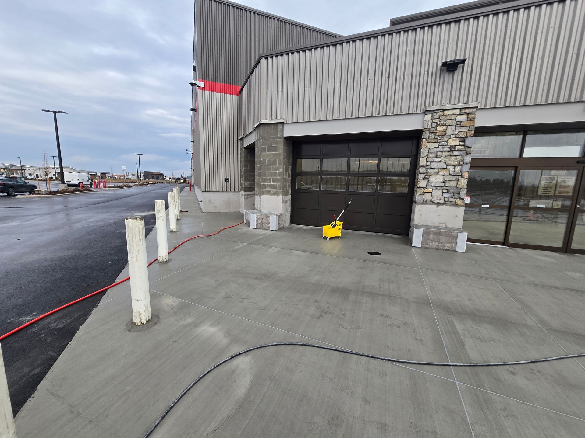 An exterior view of a modern building entrance with bollards, a concrete walkway, and a yellow mop bucket by the door.