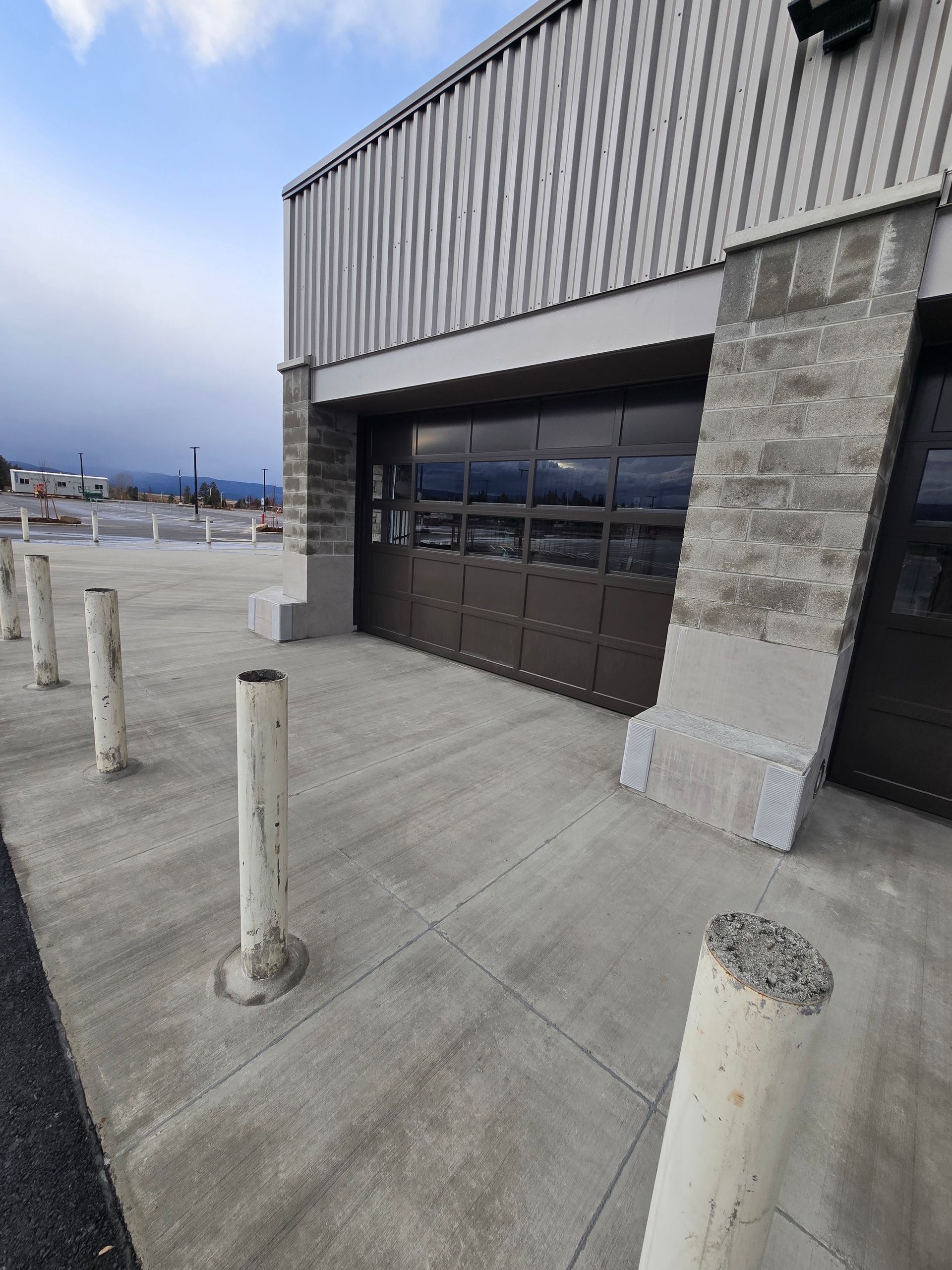A concrete exterior of a building with a dark, segmented glass garage door and several white safety bollards in front.