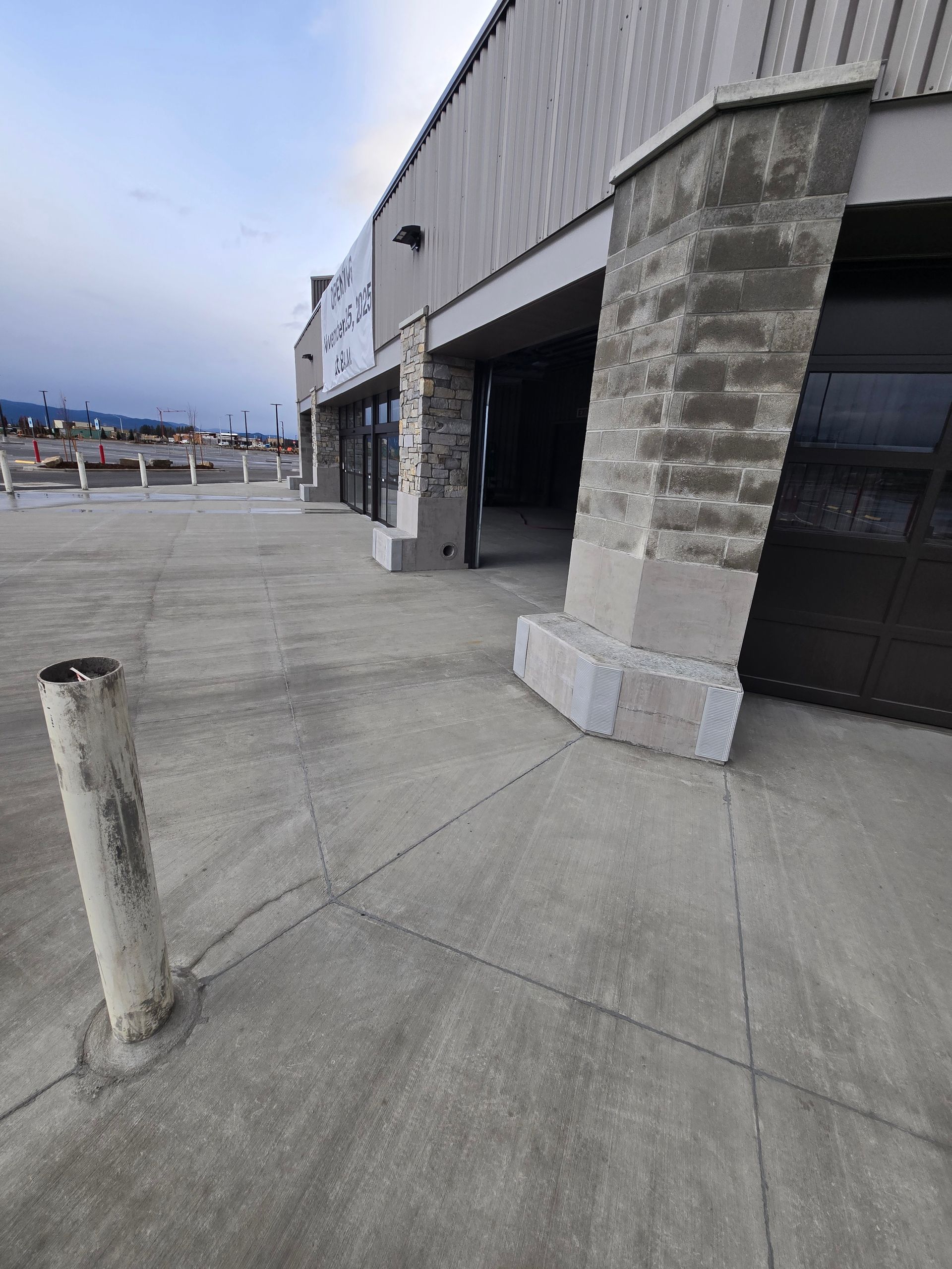 A low-angle view of a building exterior with large concrete pillars, a paved walkway, and a vertical metal safety bollard.