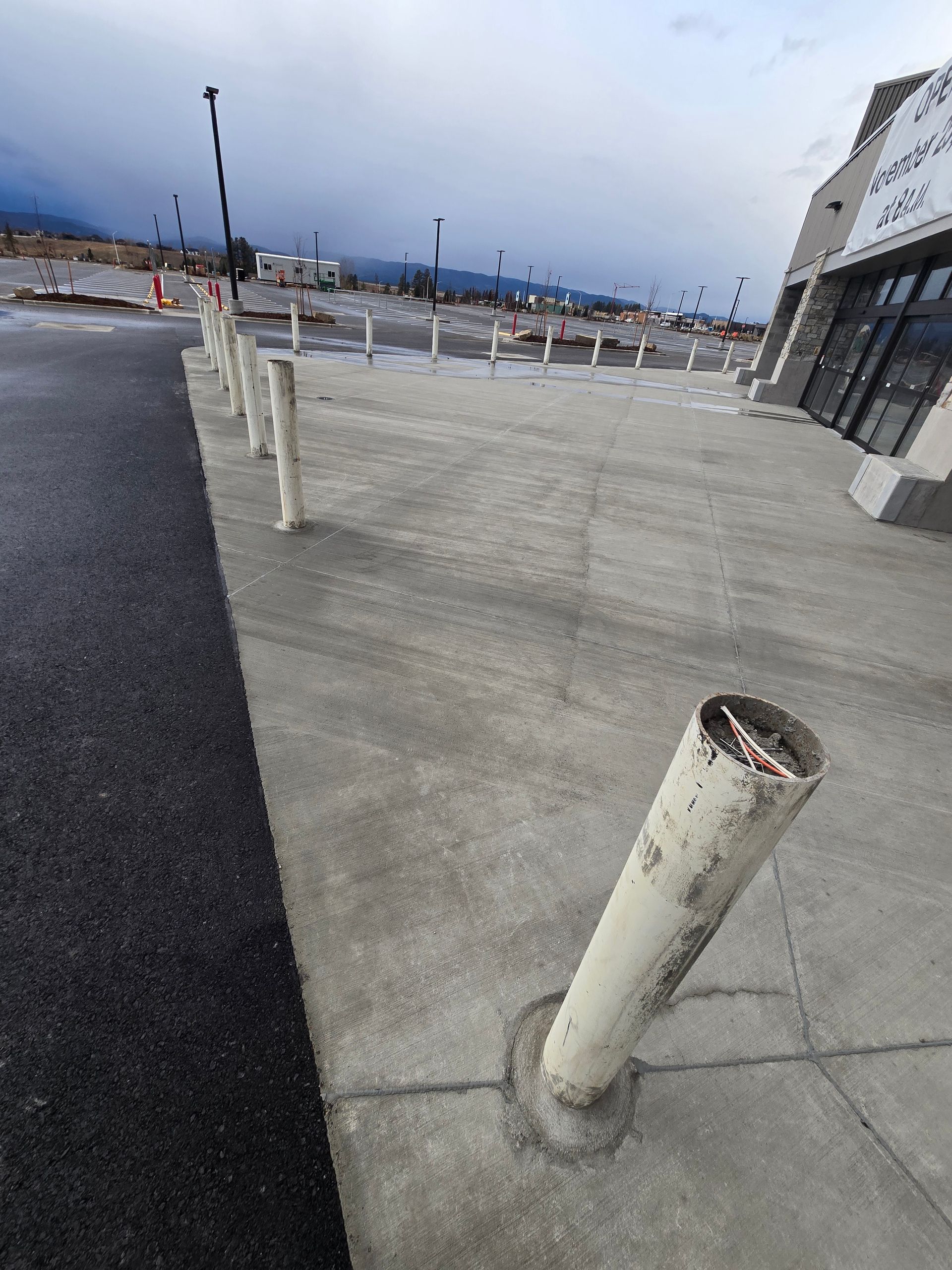 A line of white concrete bollards on a sidewalk in front of a building, with a parking lot in the background.