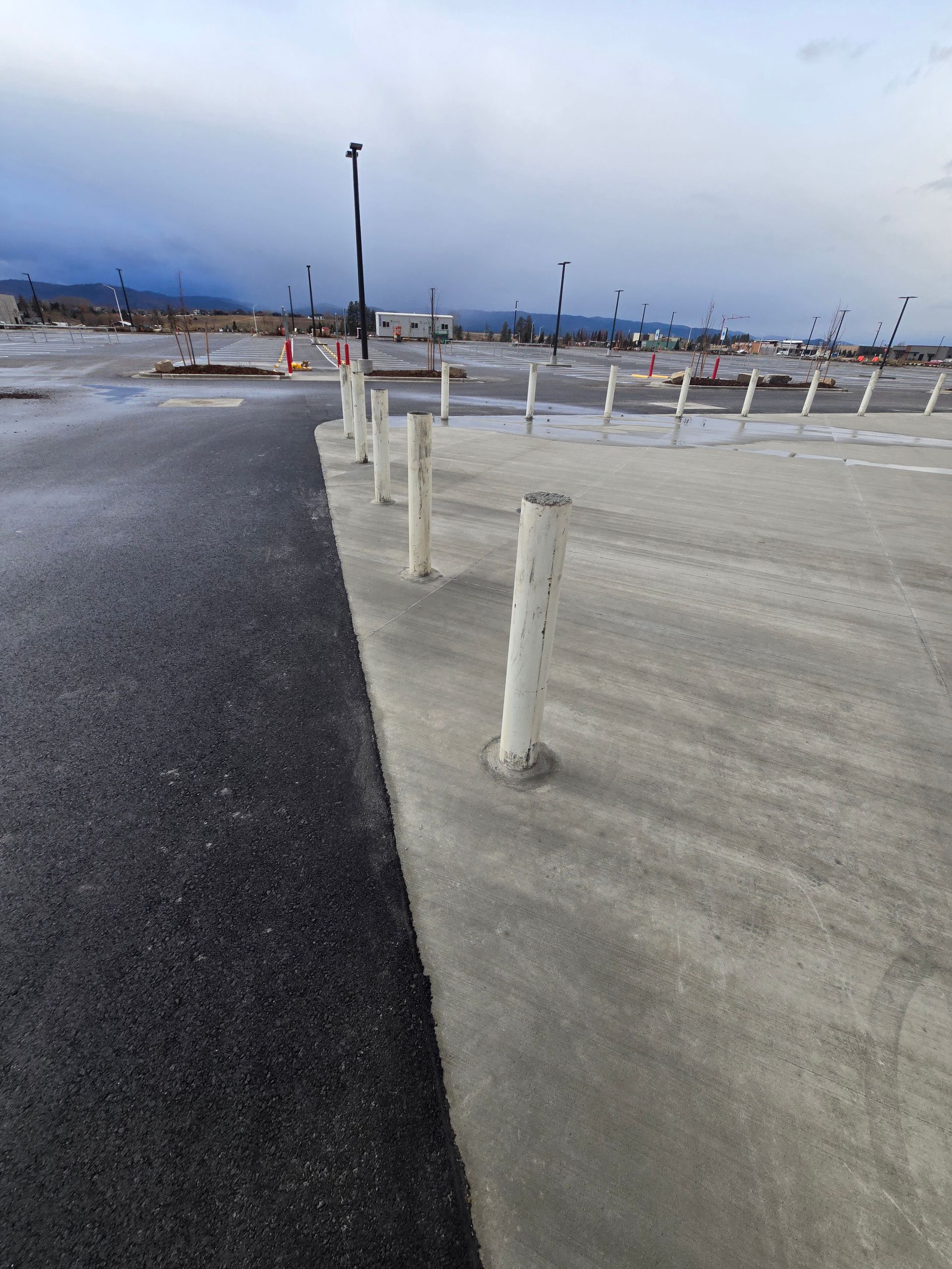 A row of white bollards separates an asphalt road from a concrete parking area under a cloudy sky.