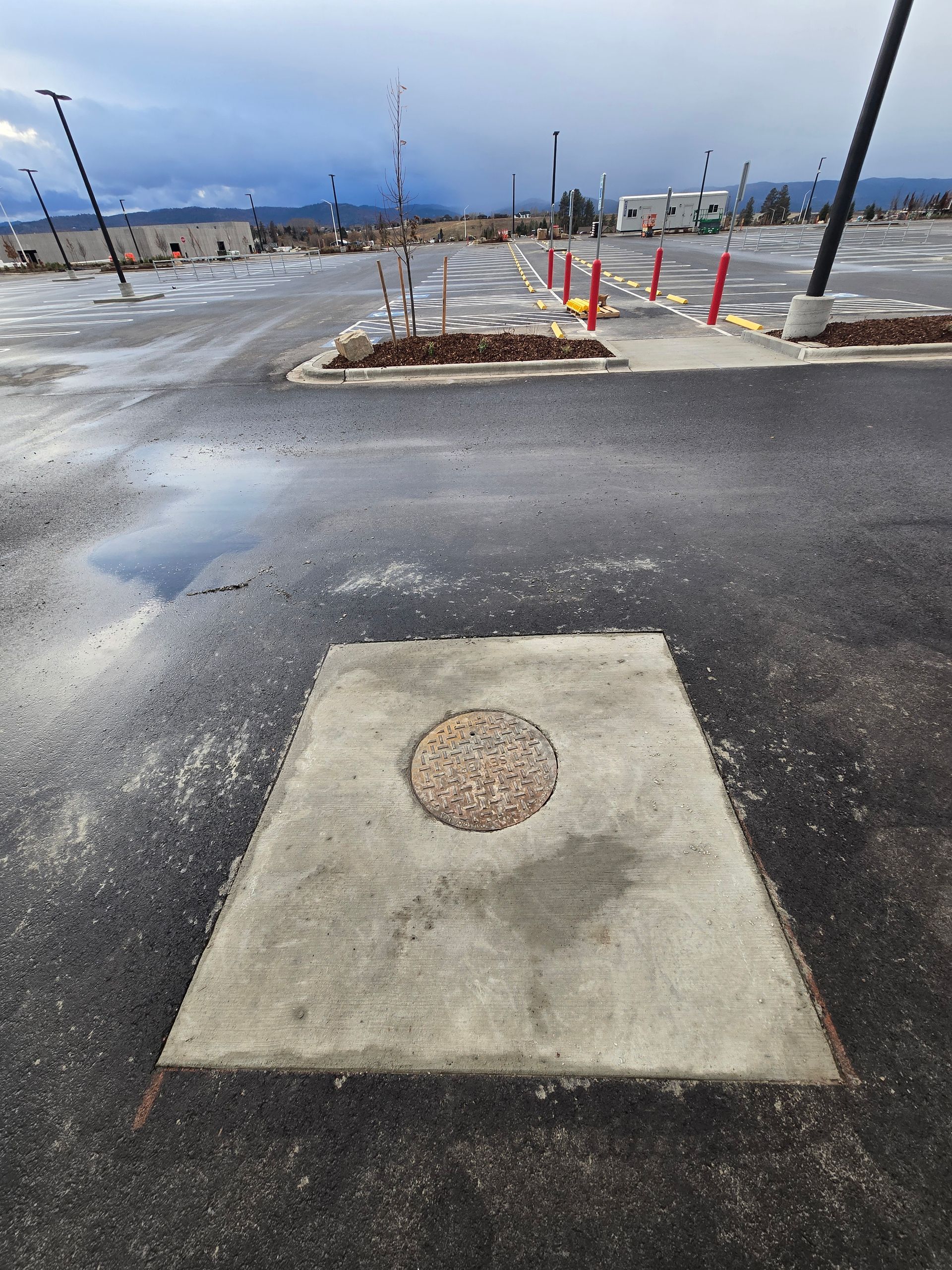 A circular utility cover centered in a rectangular concrete patch surrounded by wet asphalt in an outdoor parking lot.
