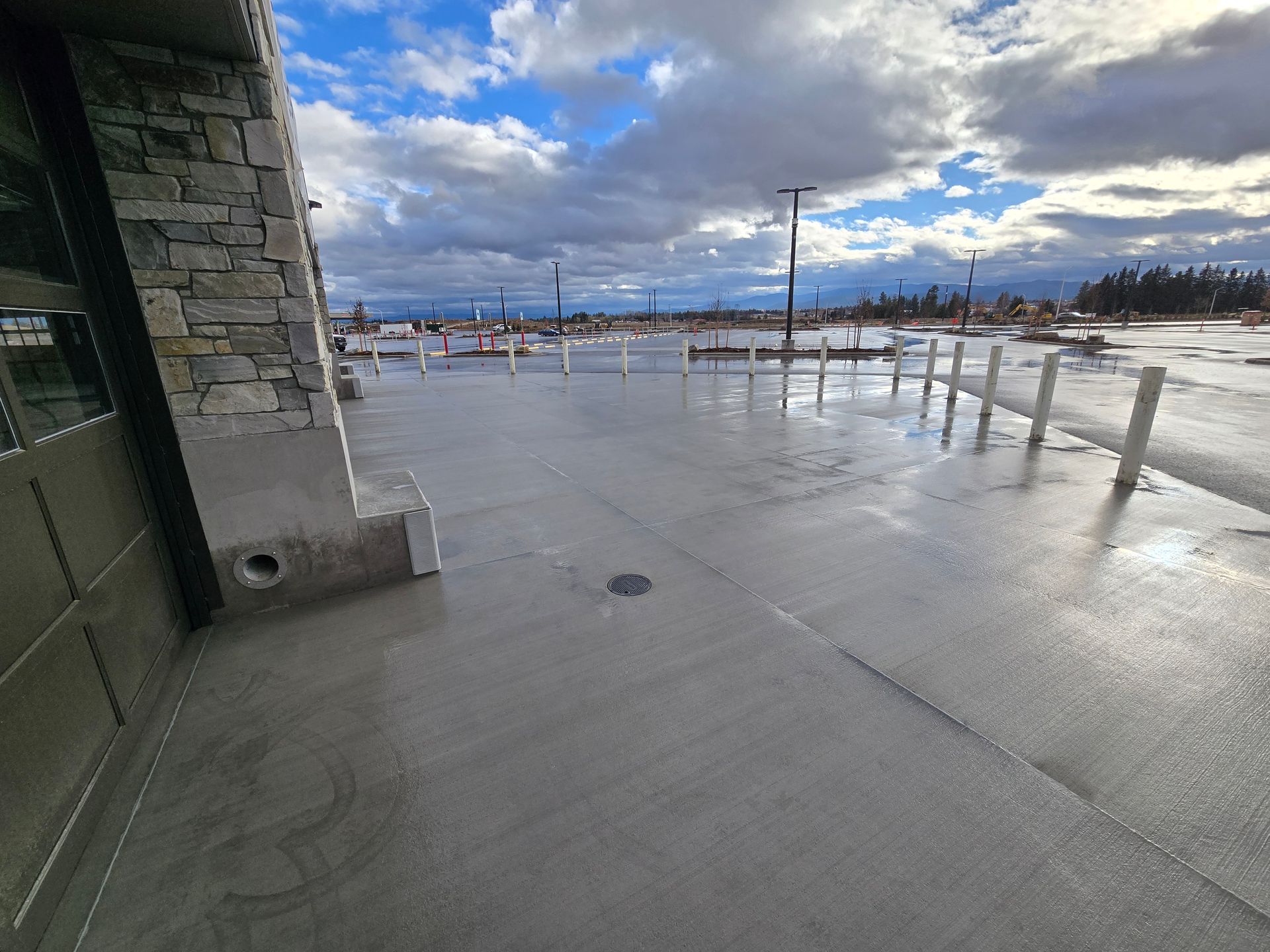 A paved outdoor area with a stone building wall on the left and a row of white bollards bordering a parking lot.