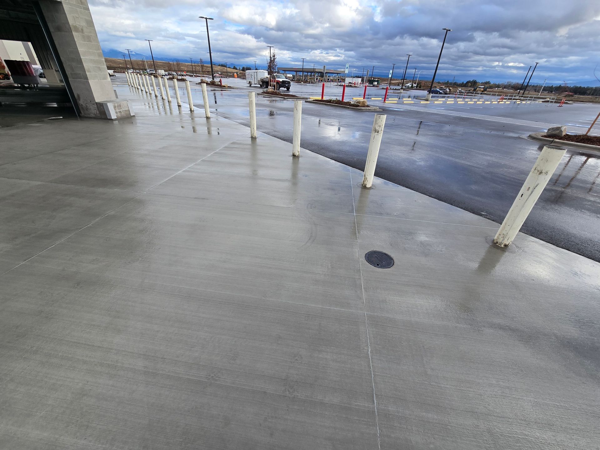 A row of white bollards lines a wet, gray concrete sidewalk next to an outdoor construction site under a cloudy sky.