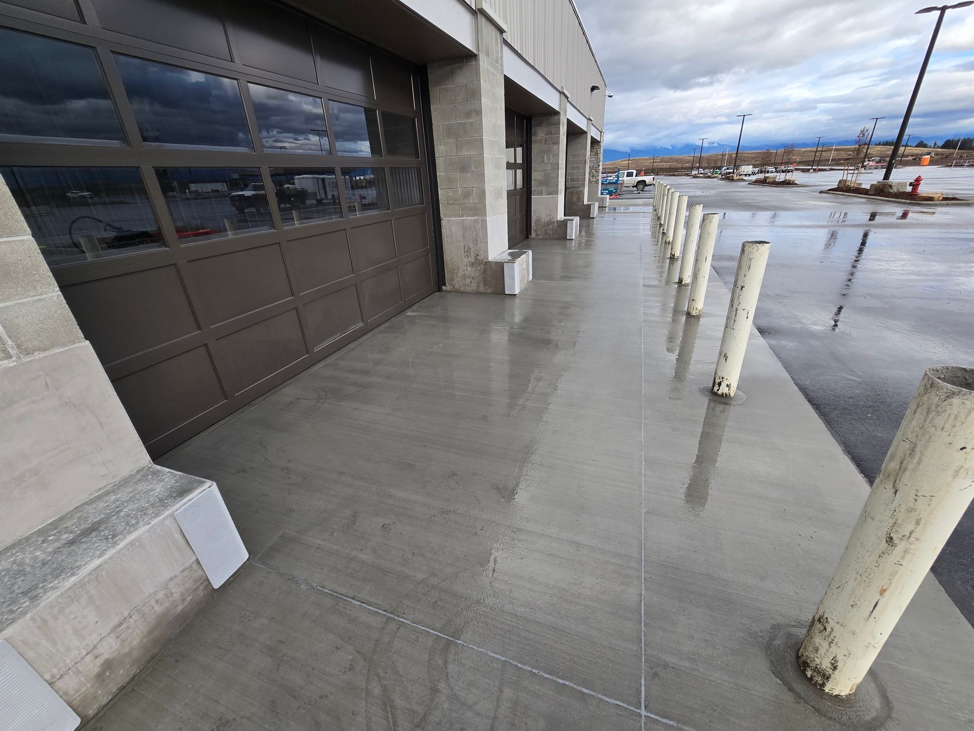 Wet concrete sidewalk outside a commercial building with glass garage doors and a row of safety bollards.