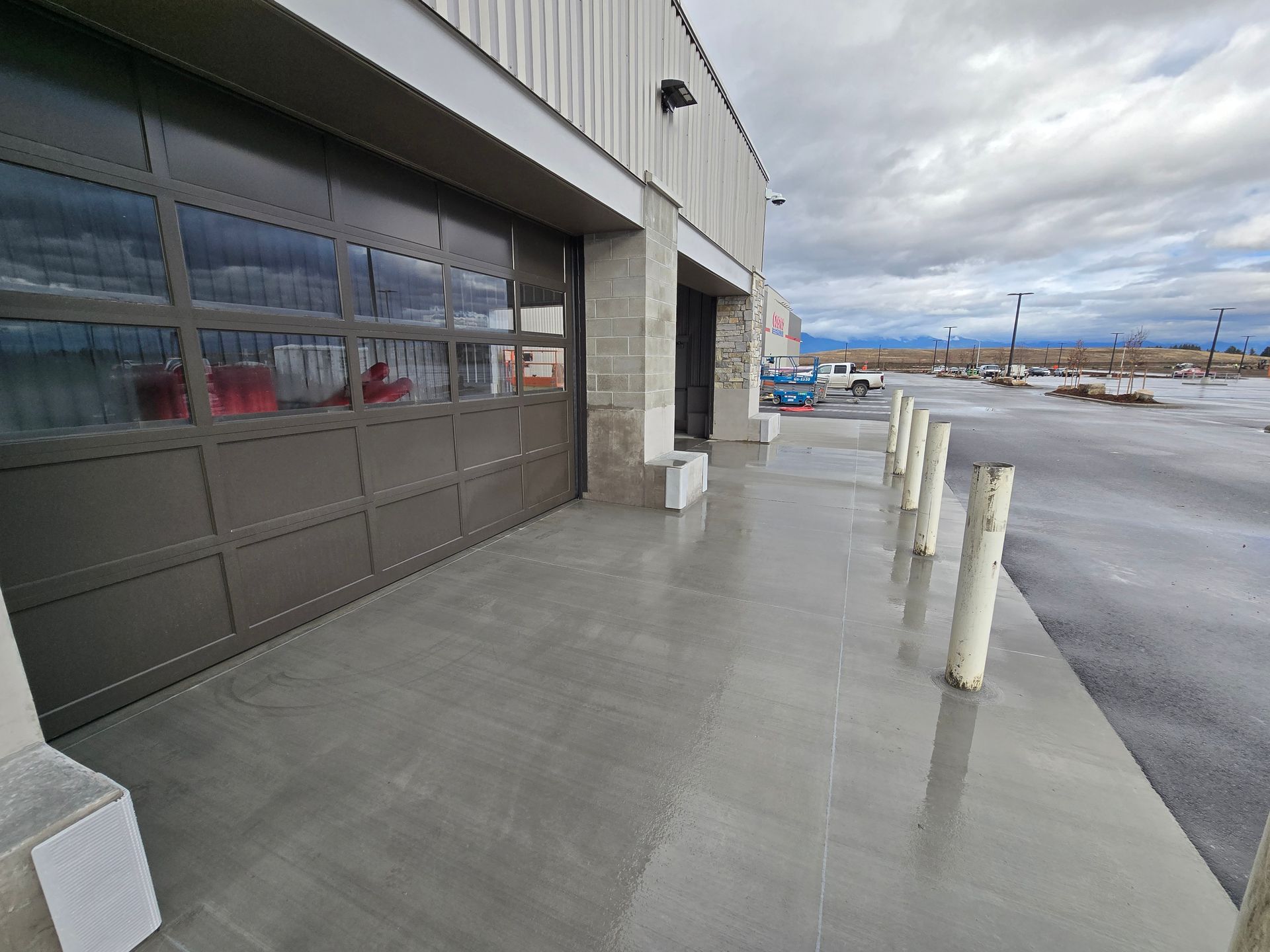 A row of white bollards lines a wet concrete walkway next to a building with a large, glass-paneled garage door.