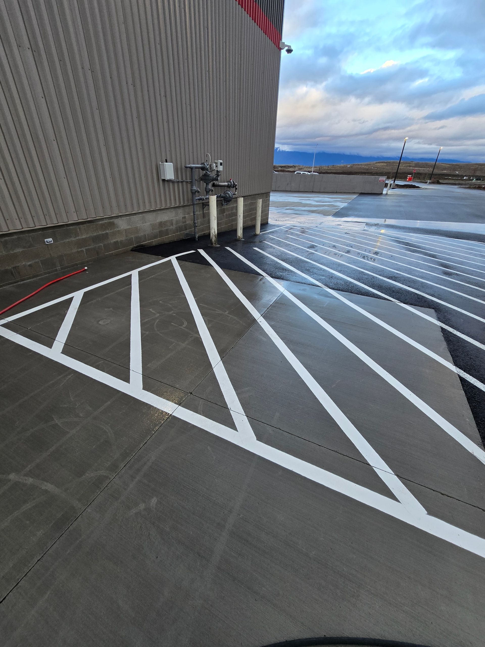 A parking lot area featuring white diagonal stripes painted on gray concrete next to a corrugated metal building wall.
