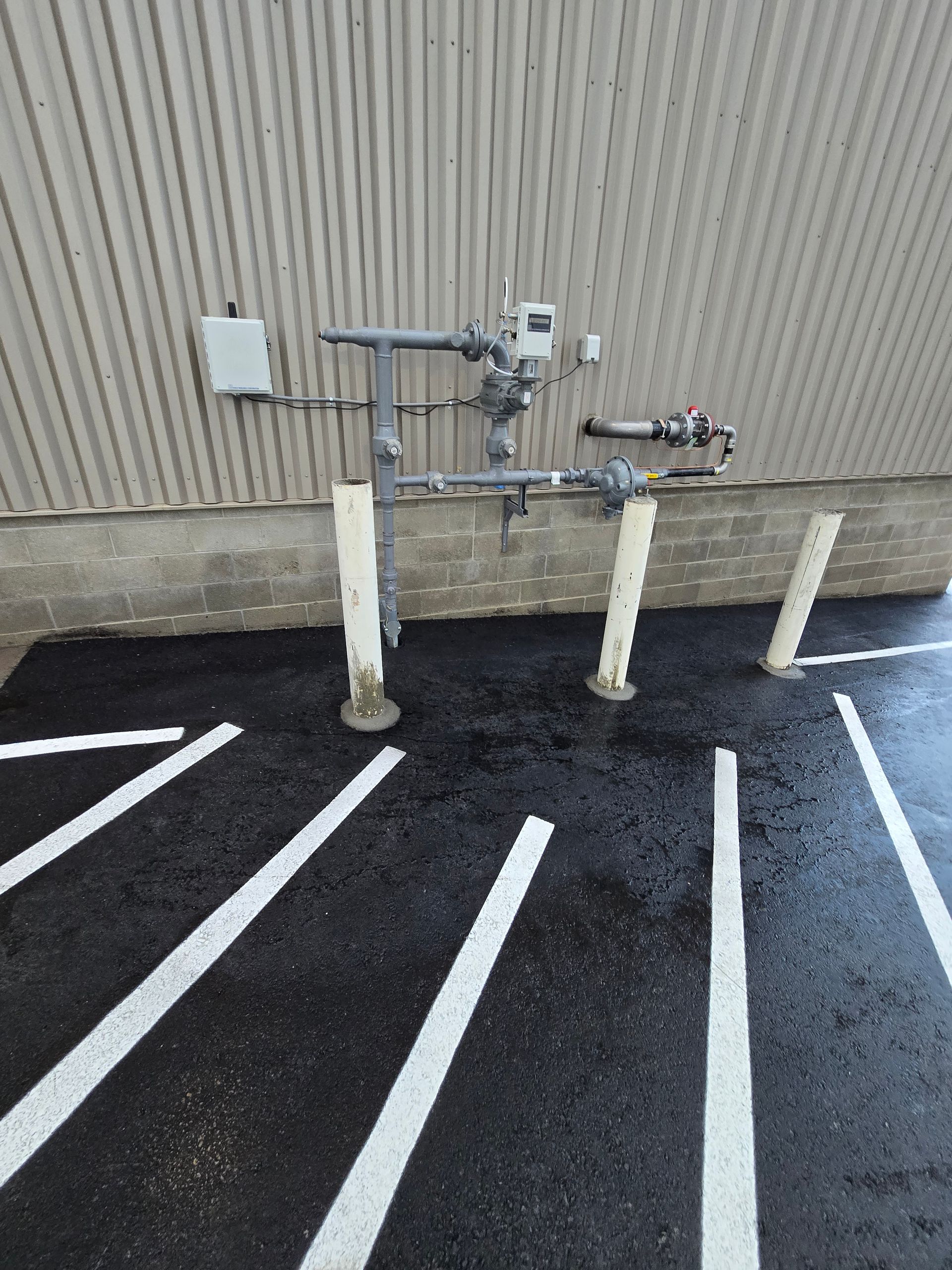 Gas meter and piping assembly mounted on a building wall above a parking lot with white-striped pavement and bollards.