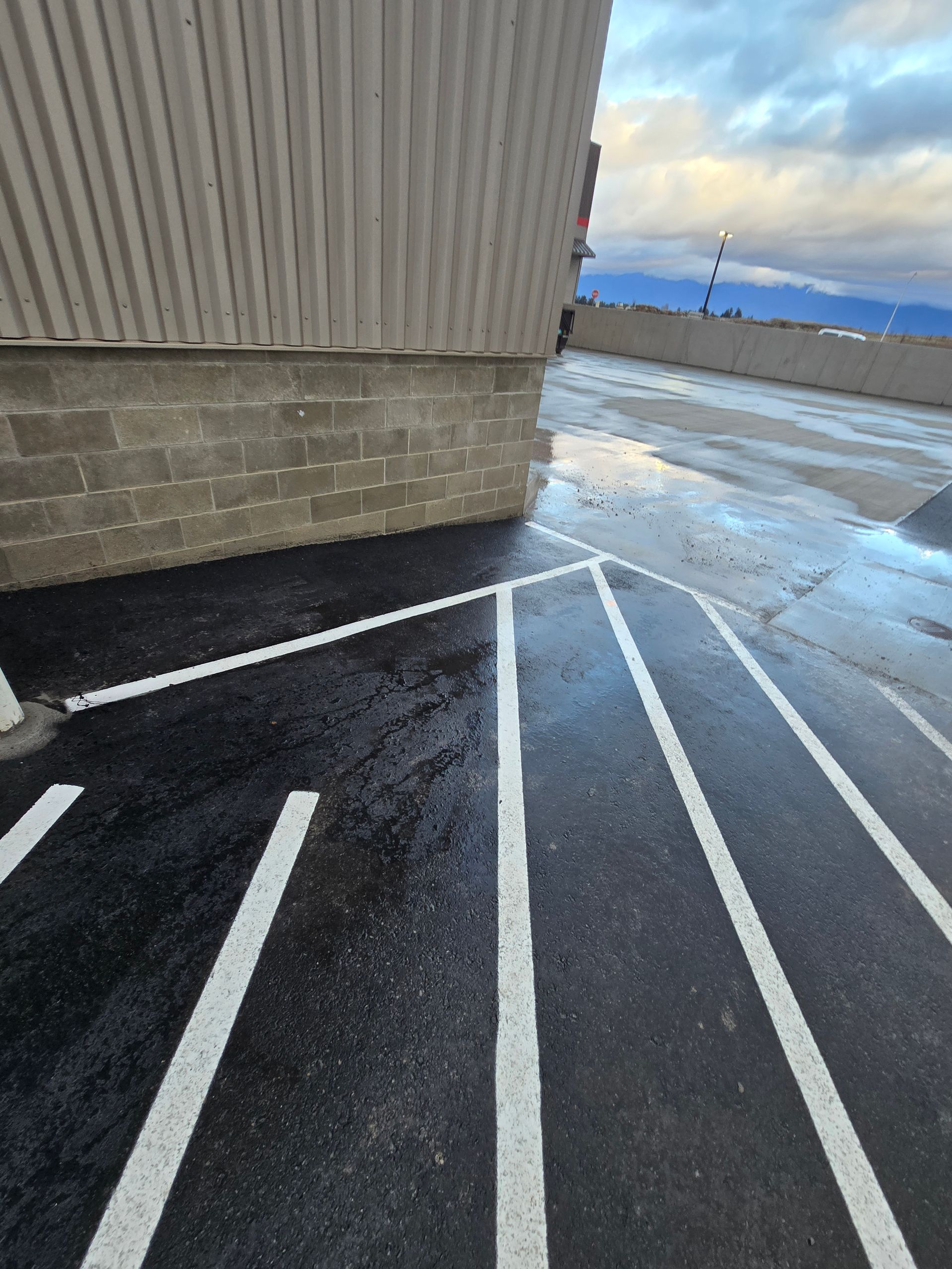 A parking lot corner showing white painted lines on black asphalt next to a building wall under a cloudy sky.