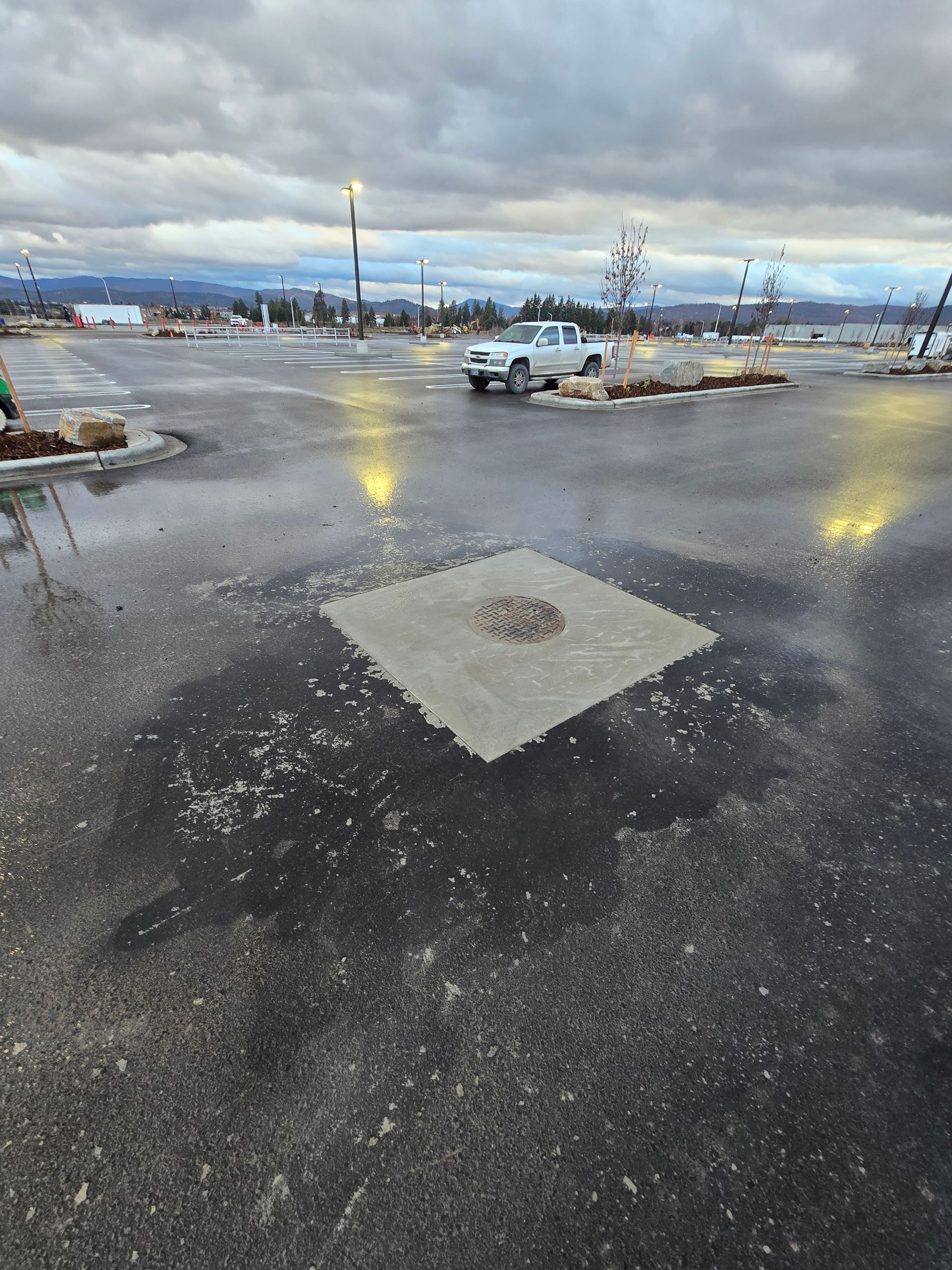 A wet, grey parking lot featuring a square concrete patch with a circular manhole cover in the center under a cloudy sky.