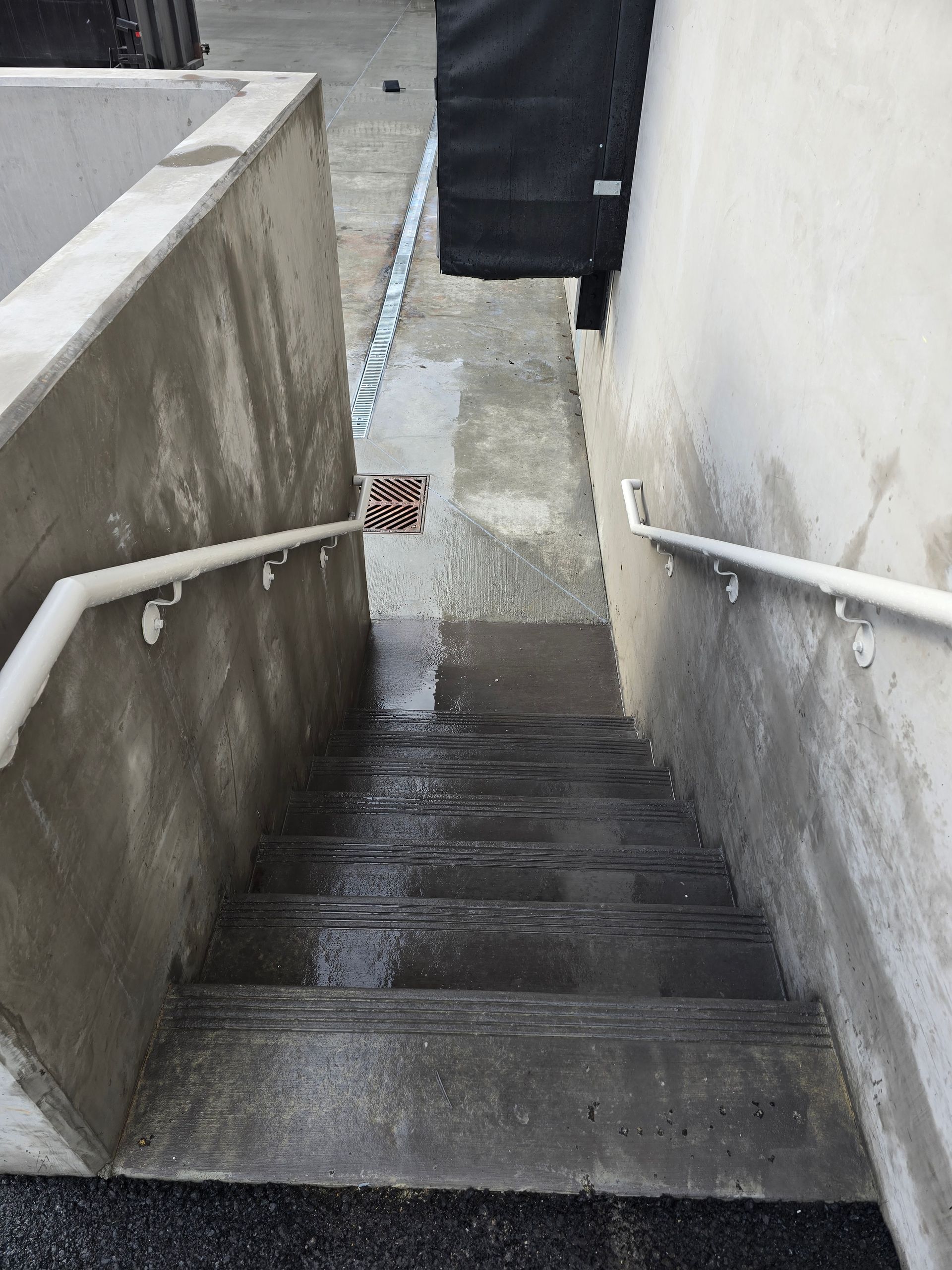 Outdoor concrete stairs with white handrails on both sides, leading down to a wet, paved area with a drain.