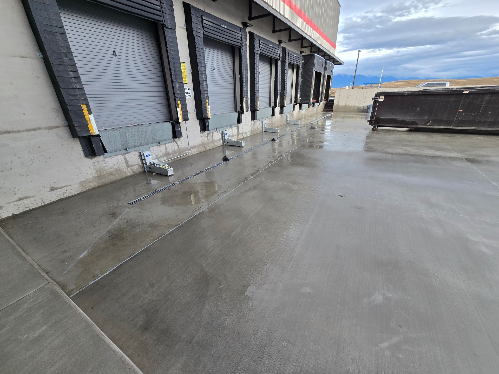 A line of loading dock doors on a concrete building exterior with a large dumpster on the right.