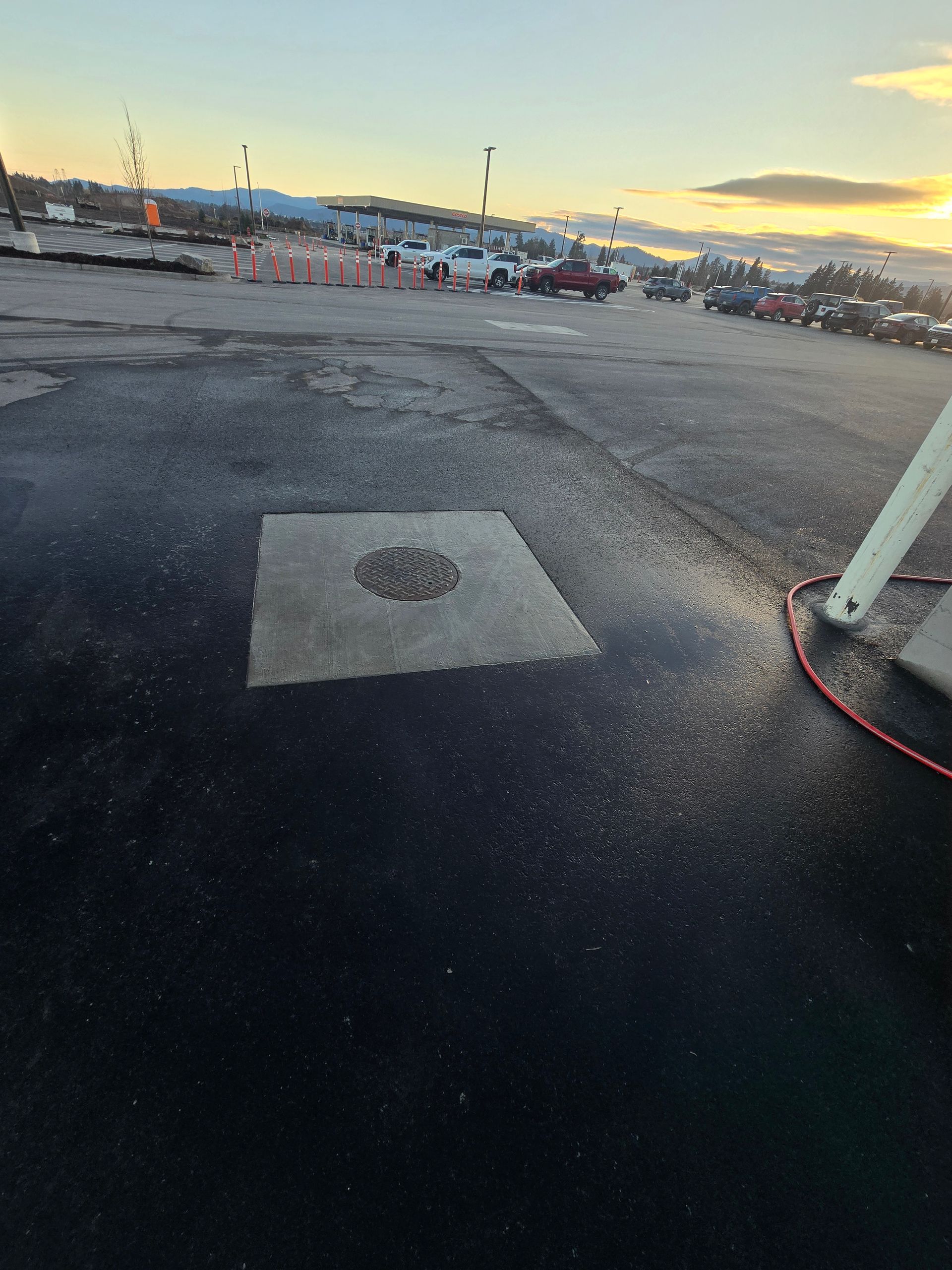 A square concrete patch with a round access cover in the middle of a black asphalt gas station lot at sunset.