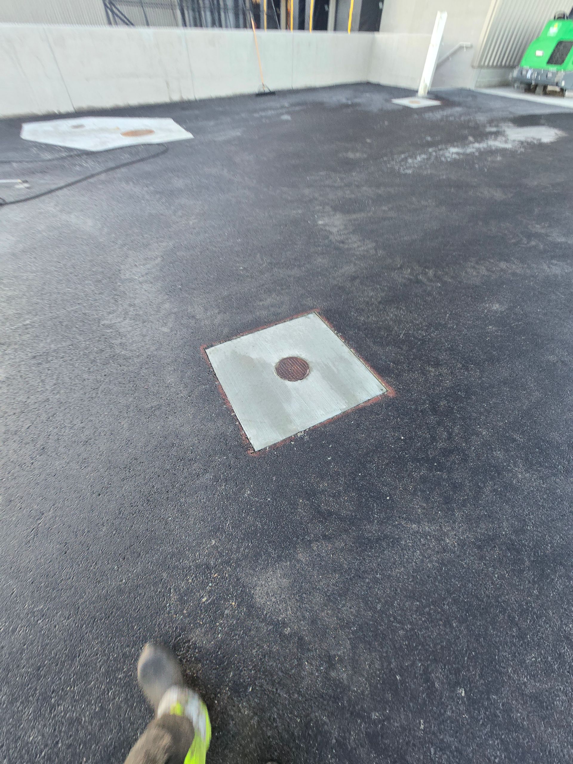 An asphalt parking lot featuring two concrete patches with circular access covers and a green object in the background.