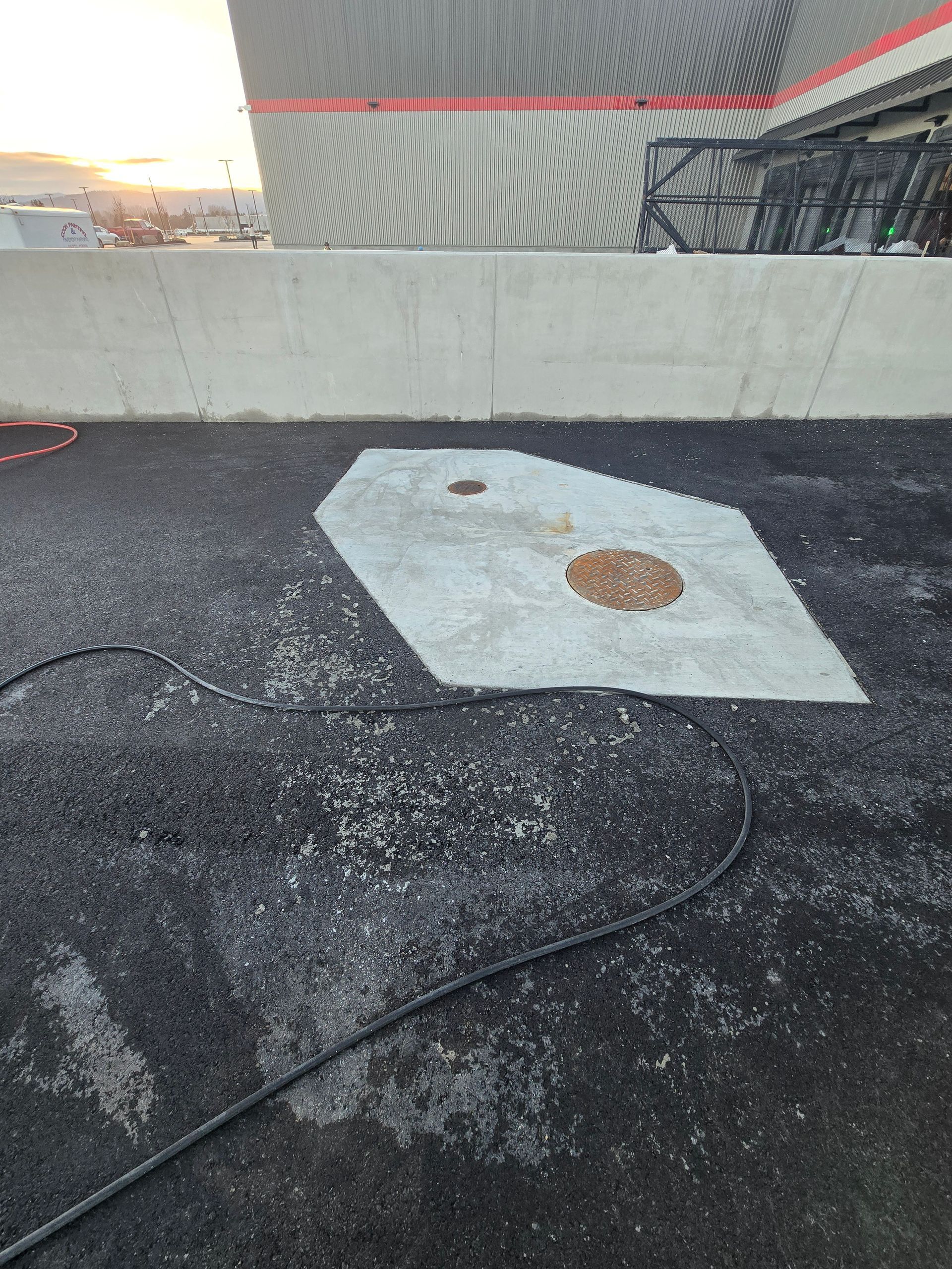 A concrete patch with two circular utility access covers sits in the middle of a dark asphalt parking lot at sunset.