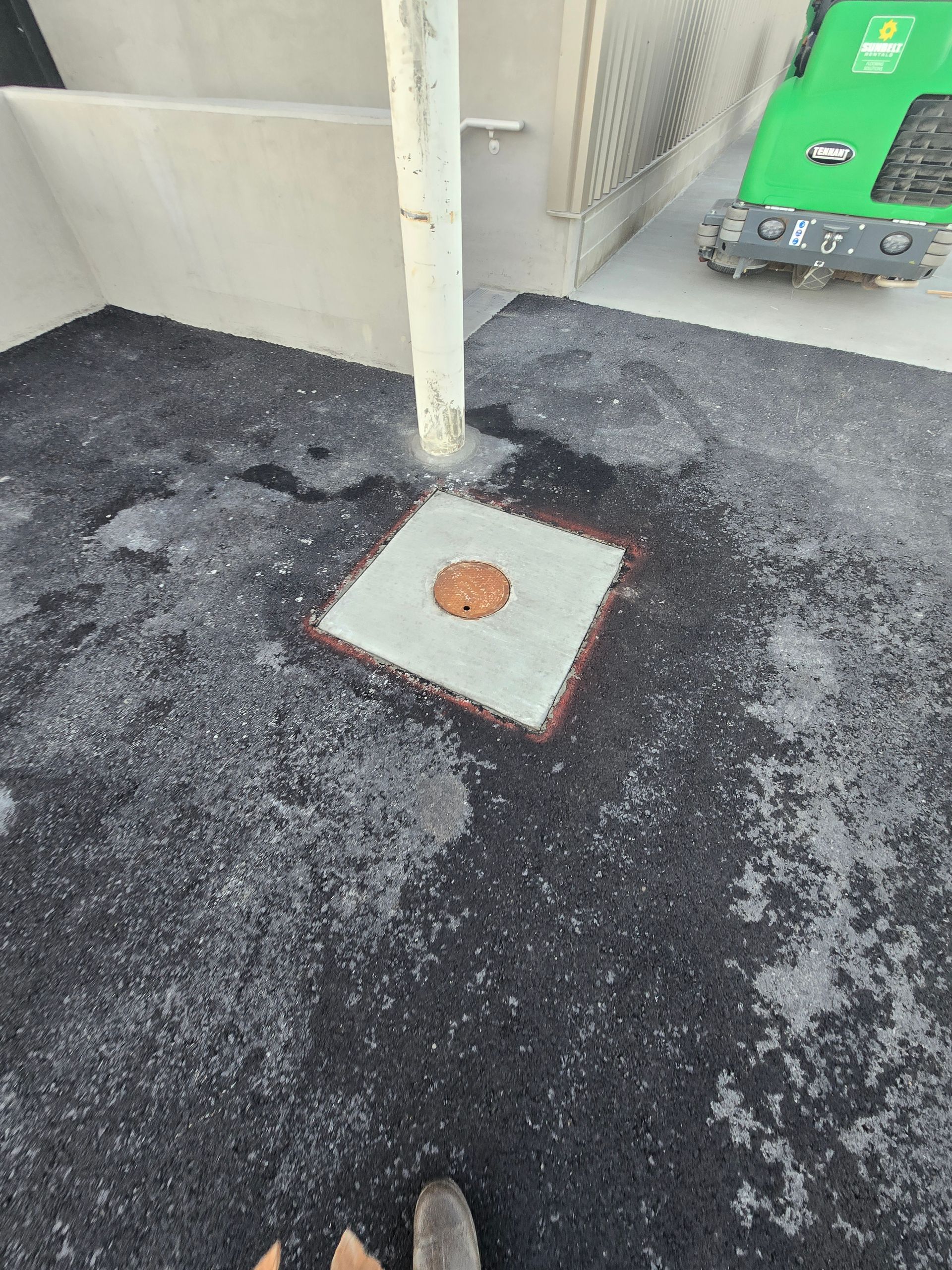 A square concrete access hatch with a circular center plate set in newly laid black asphalt near a wall and white pole.