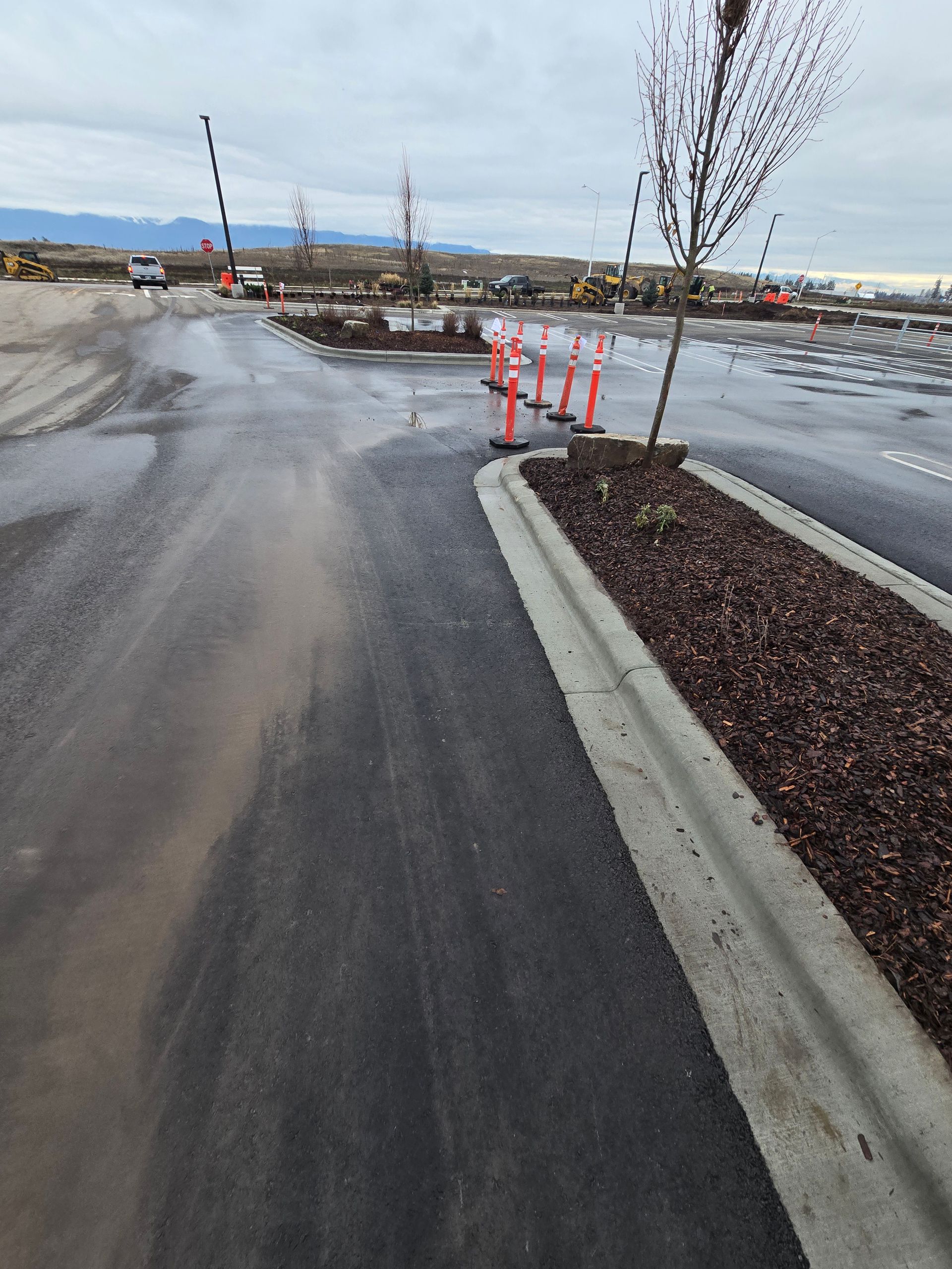 A construction site parking lot with wet asphalt, a mulch-filled median, and a line of orange safety cones.