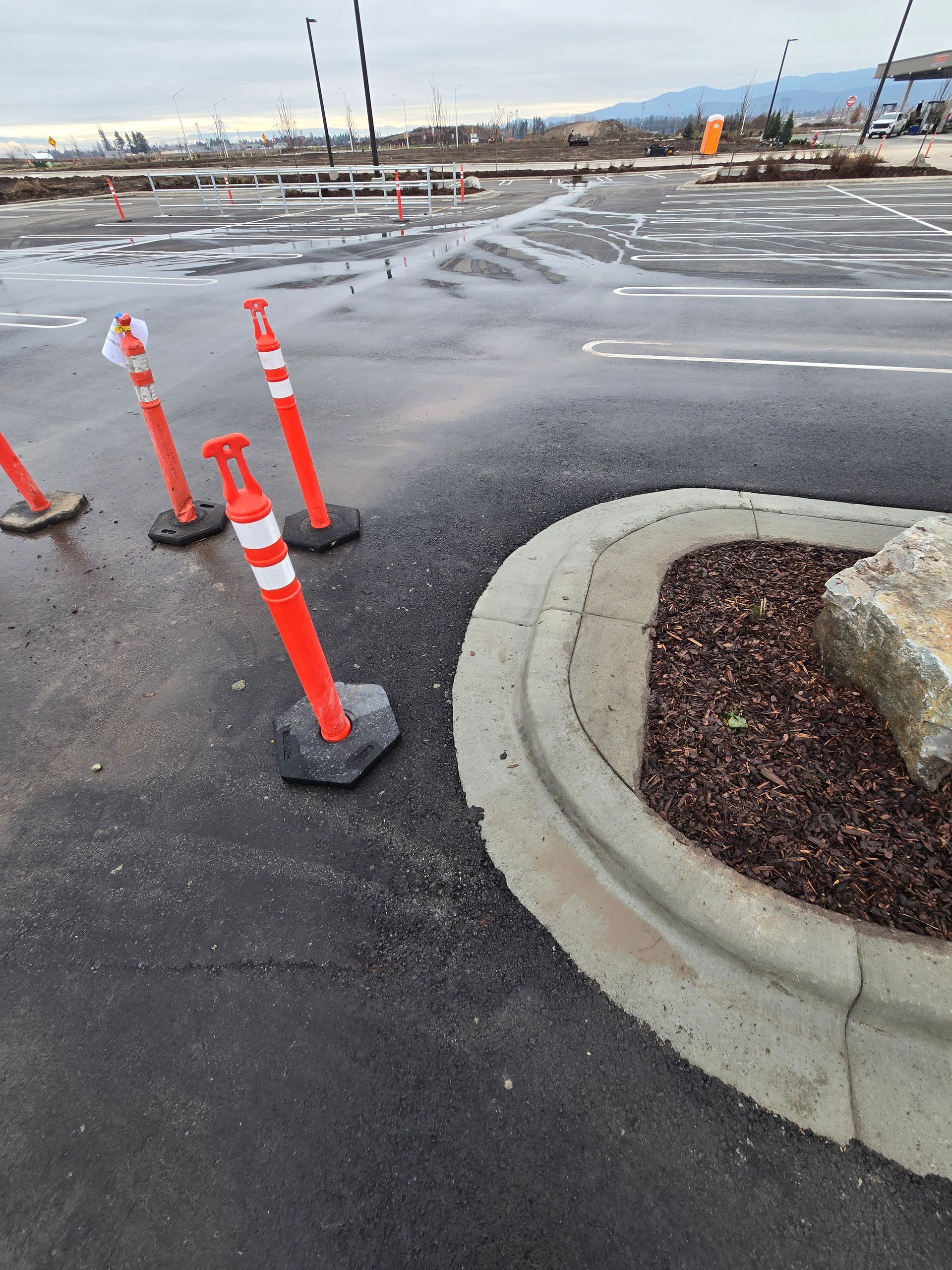 Three orange traffic delineators stand on a wet asphalt parking lot near a curved concrete curb and a rock planter.