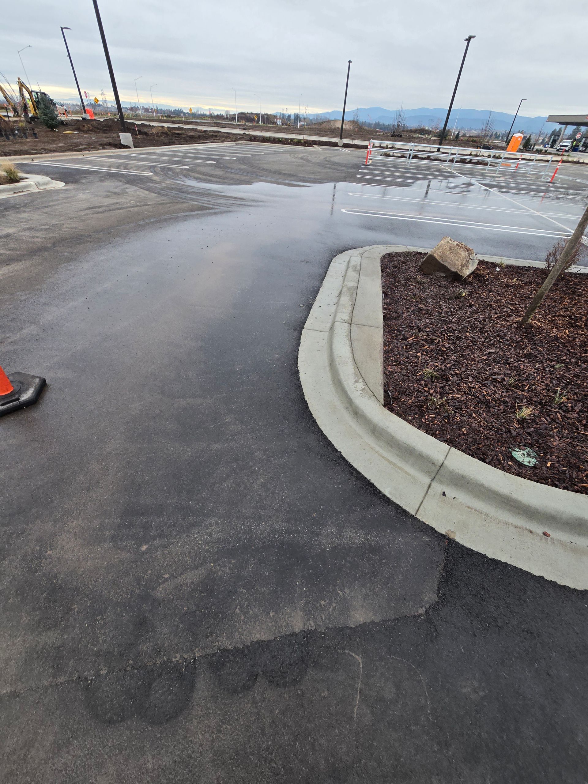 A parking lot under construction with standing water near a curved concrete curb and dark mulch landscaping.