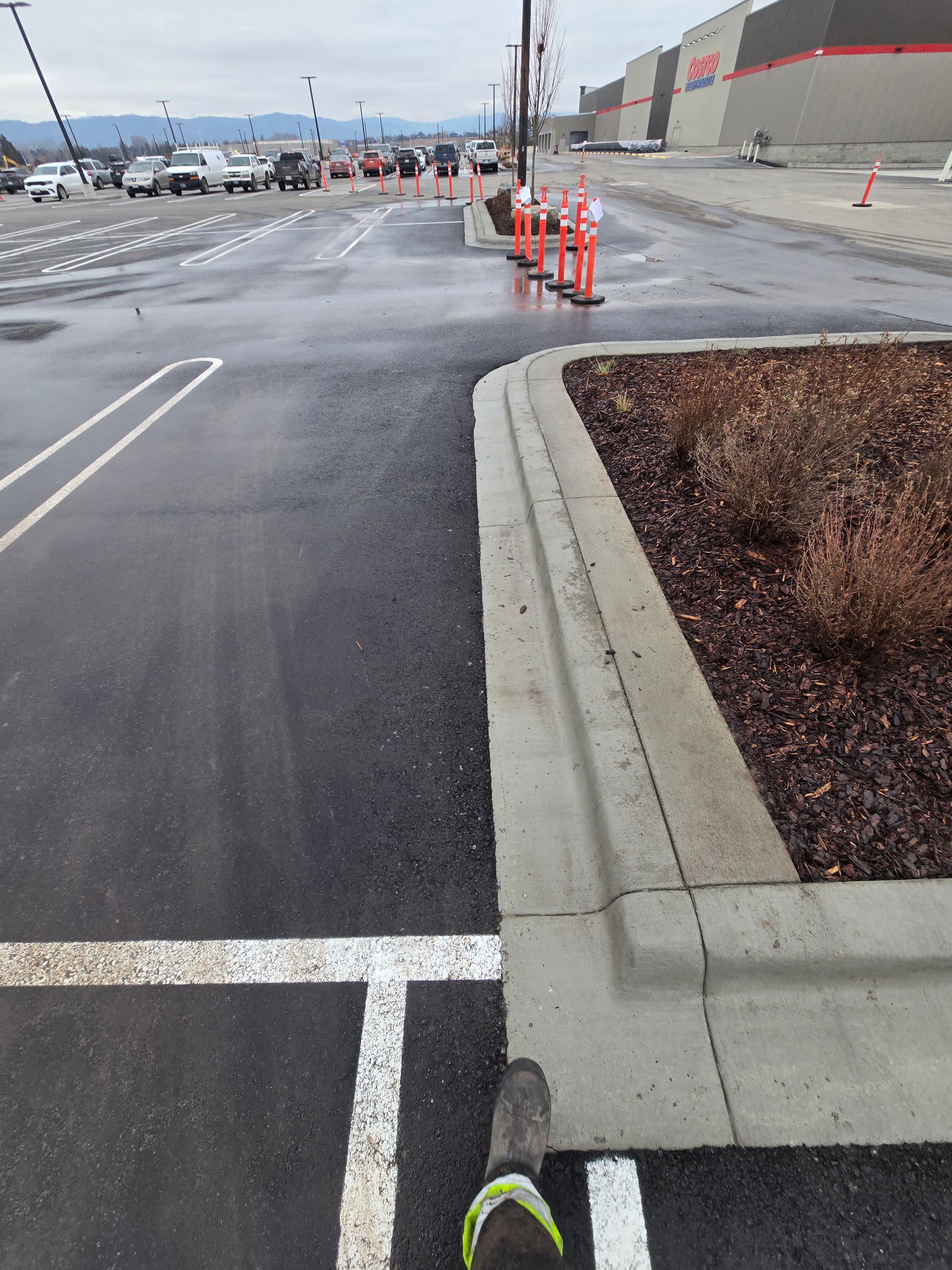 A first-person view of a person standing at a concrete curb in a parking lot, with orange traffic cones in the distance.