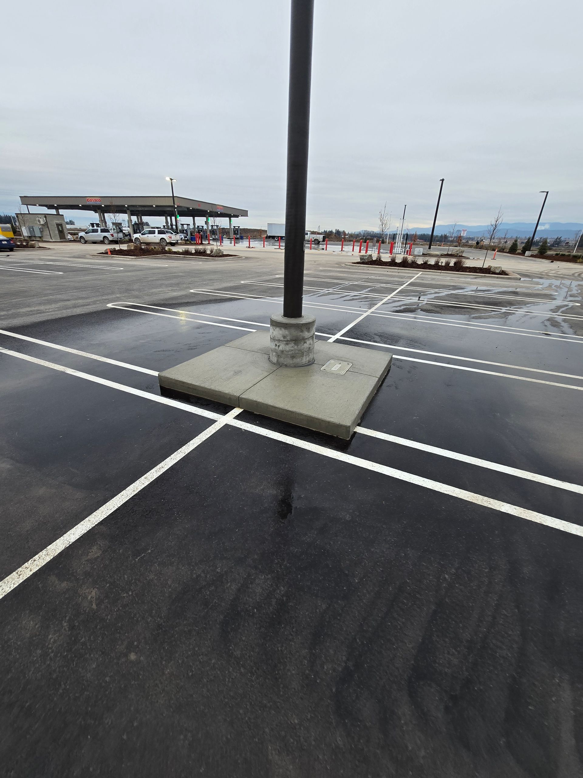 A light pole base centered in an asphalt parking lot, with a gas station canopy and construction equipment in the distance.