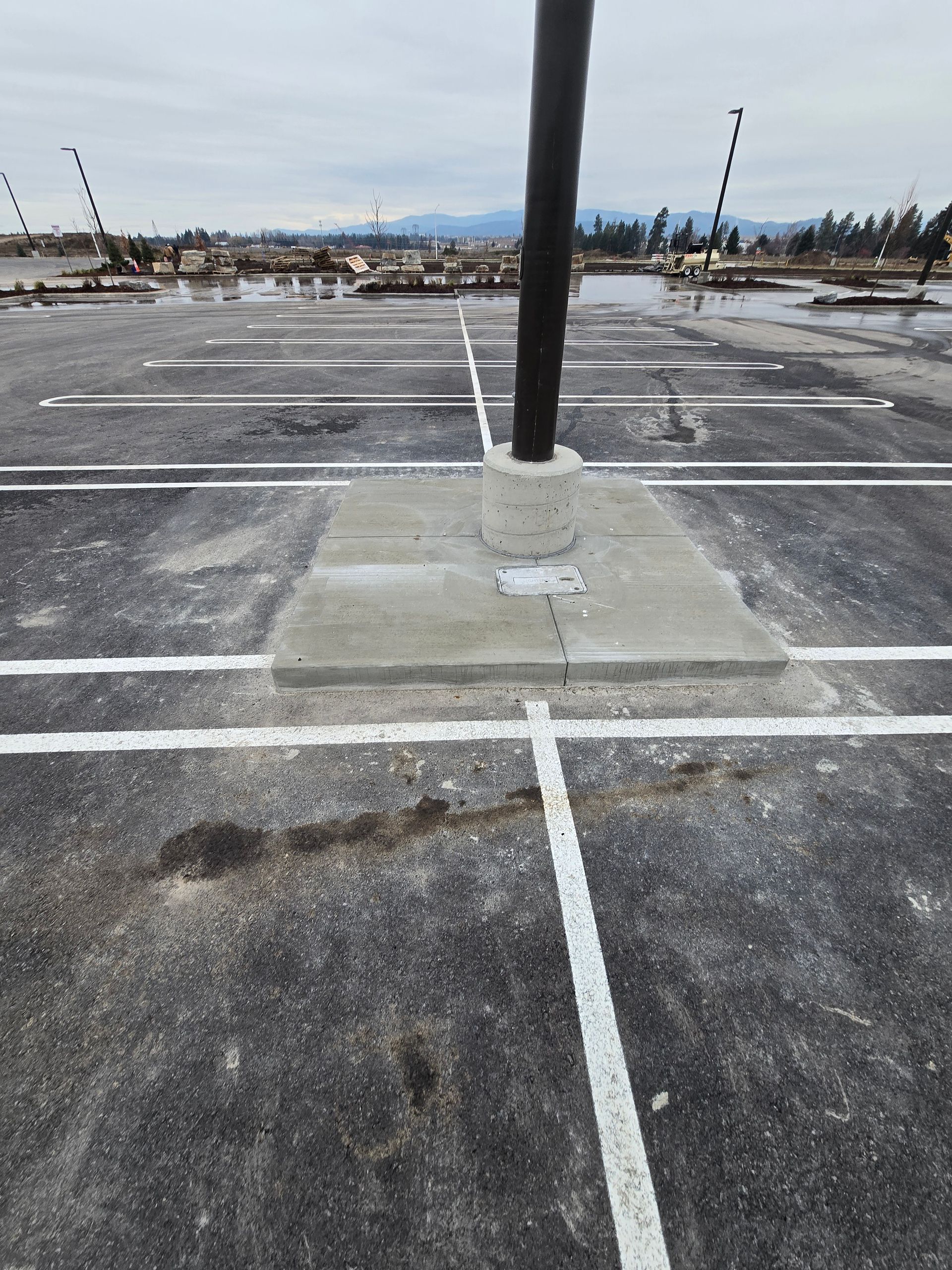 A light pole stands on a square concrete base in a paved parking lot with painted white parking space lines.