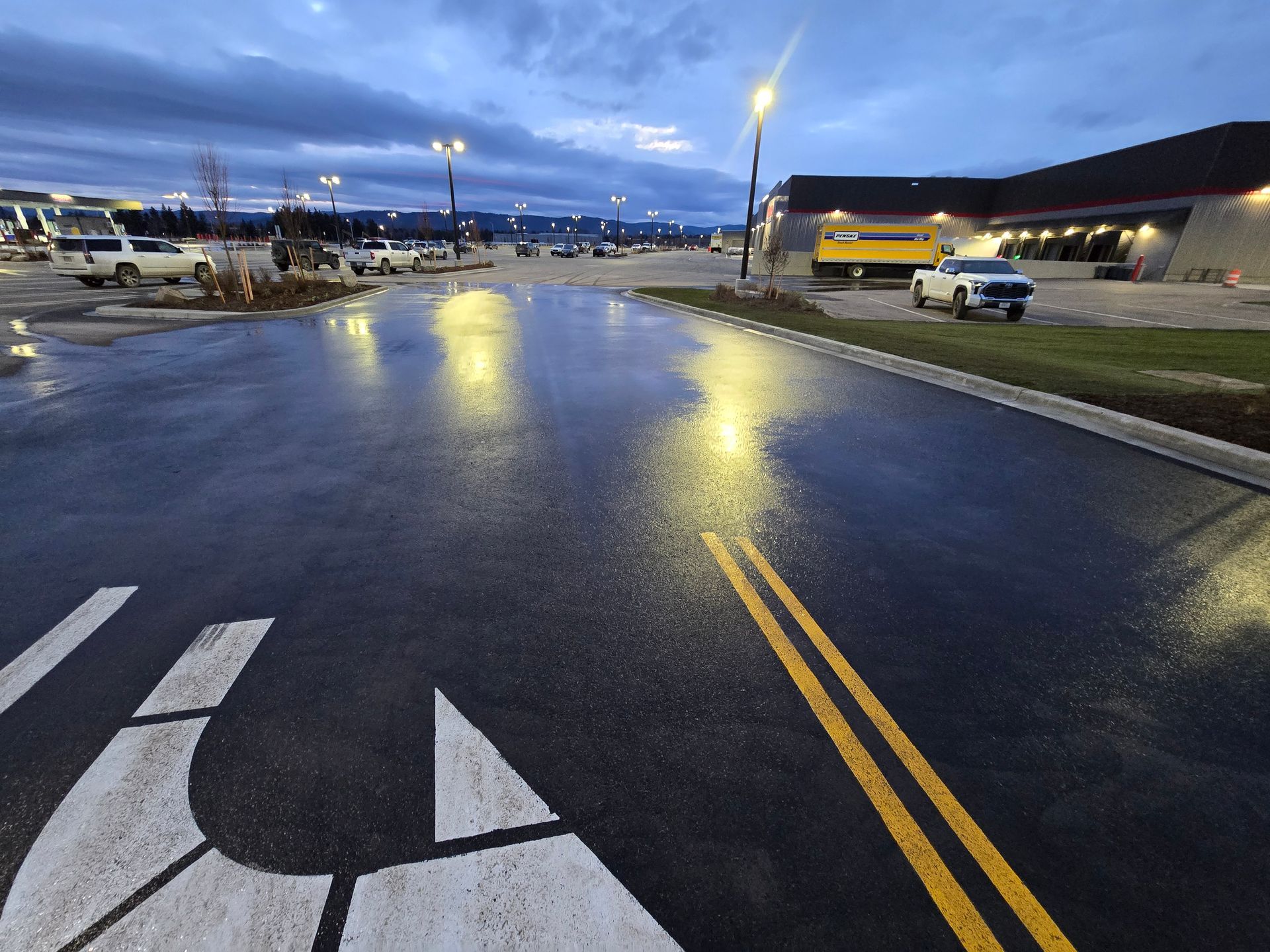 A wet parking lot at dusk, with yellow lane lines, a white arrow painted on the pavement, and distant building lights.
