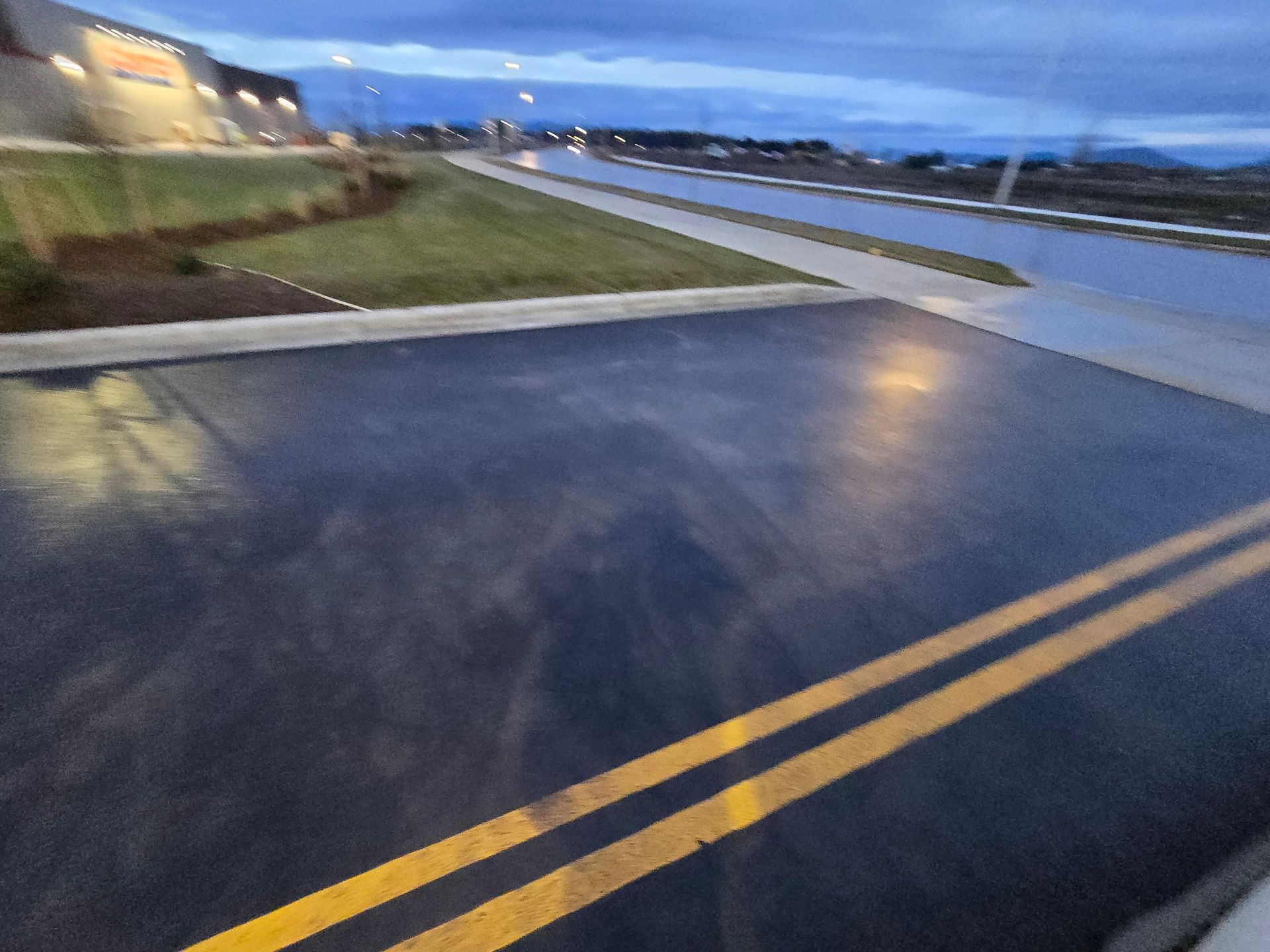 A freshly paved, dark asphalt surface with double yellow center lines, set near a building and road at dusk.