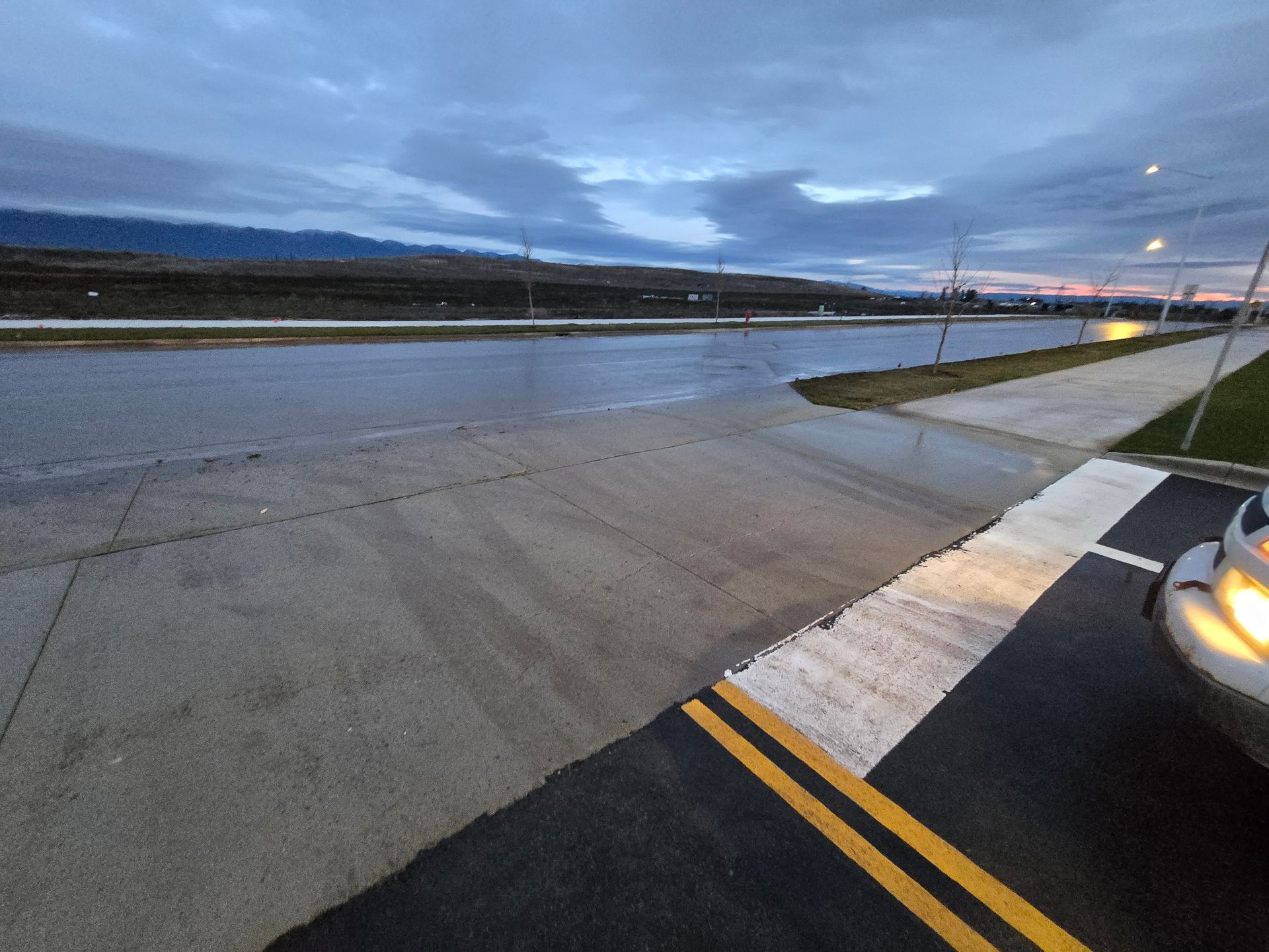 A paved road at dusk with a crosswalk, looking out toward a distant, low-lying hillside under a cloudy sky.