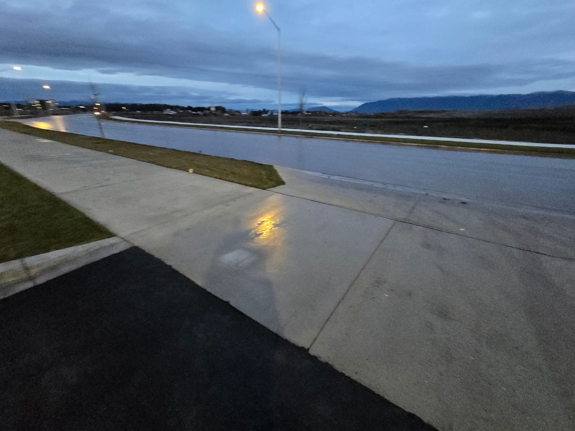 A concrete sidewalk and street at twilight with pooled water reflecting a streetlamp and a distant mountain range.