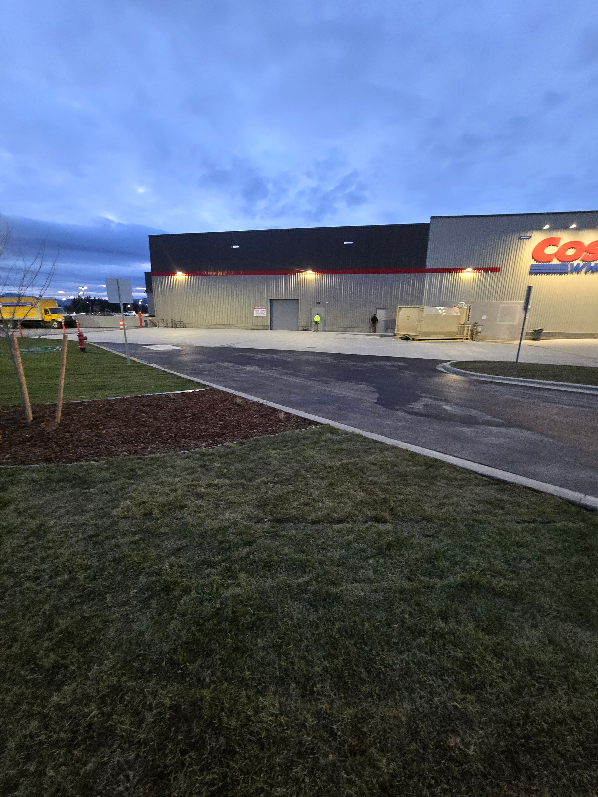 A Costco warehouse exterior during twilight, with a grassy foreground and a paved parking area.