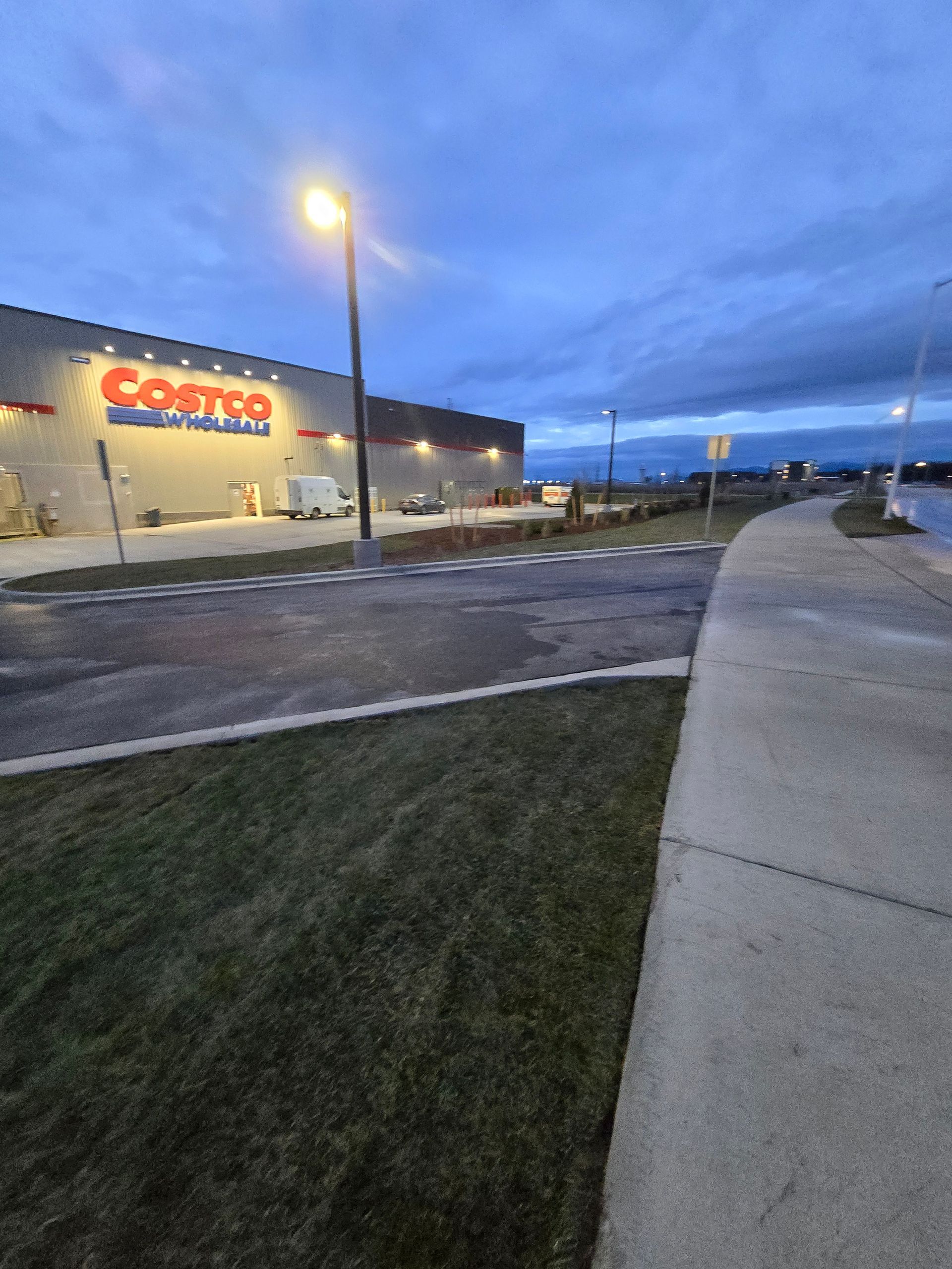Exterior view of a Costco warehouse at dusk, featuring a lit parking lot, a grassy area, and a sidewalk in the foreground.