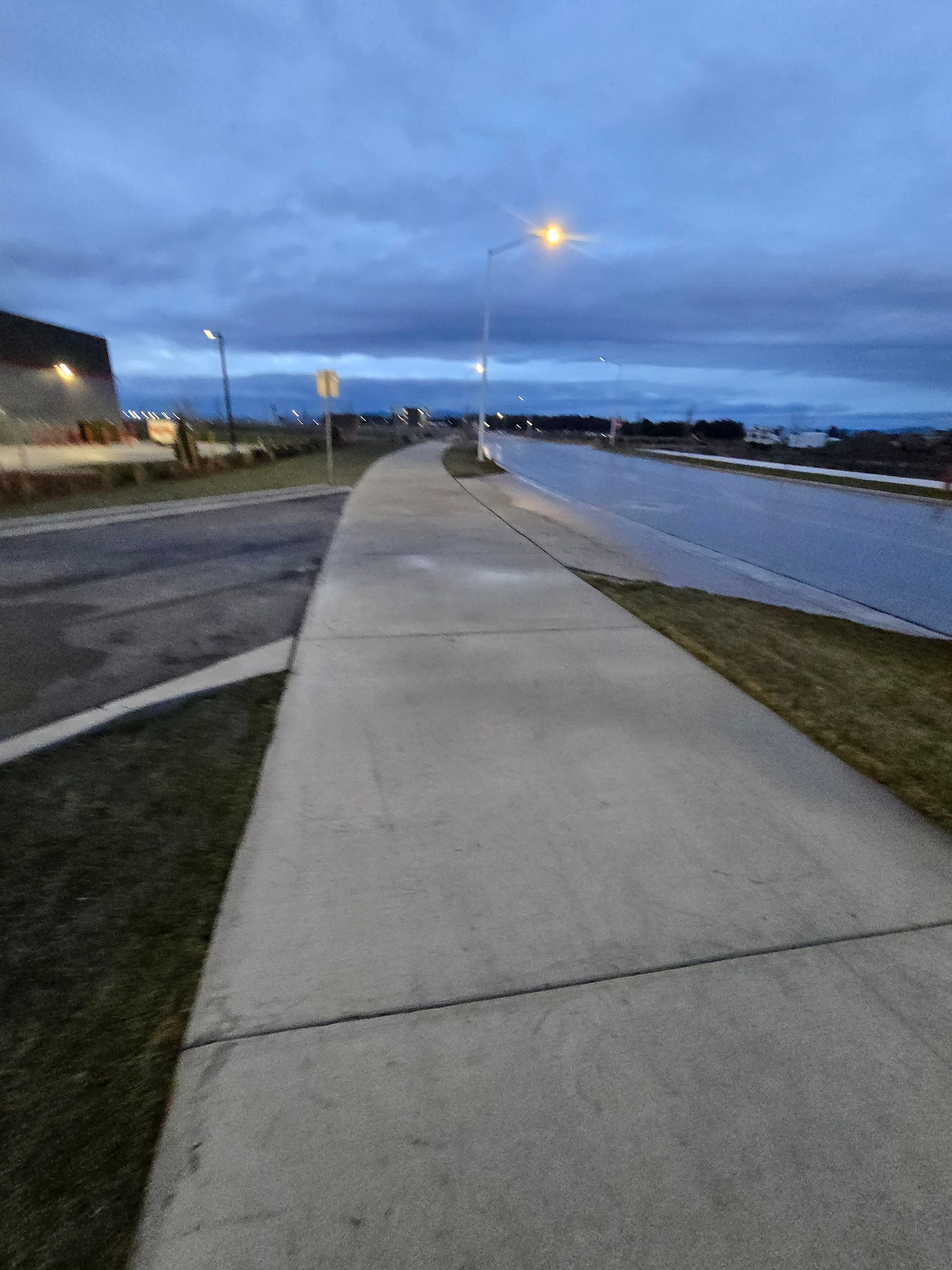 A concrete sidewalk stretches into the distance at dusk, running parallel to a road under a cloudy sky.