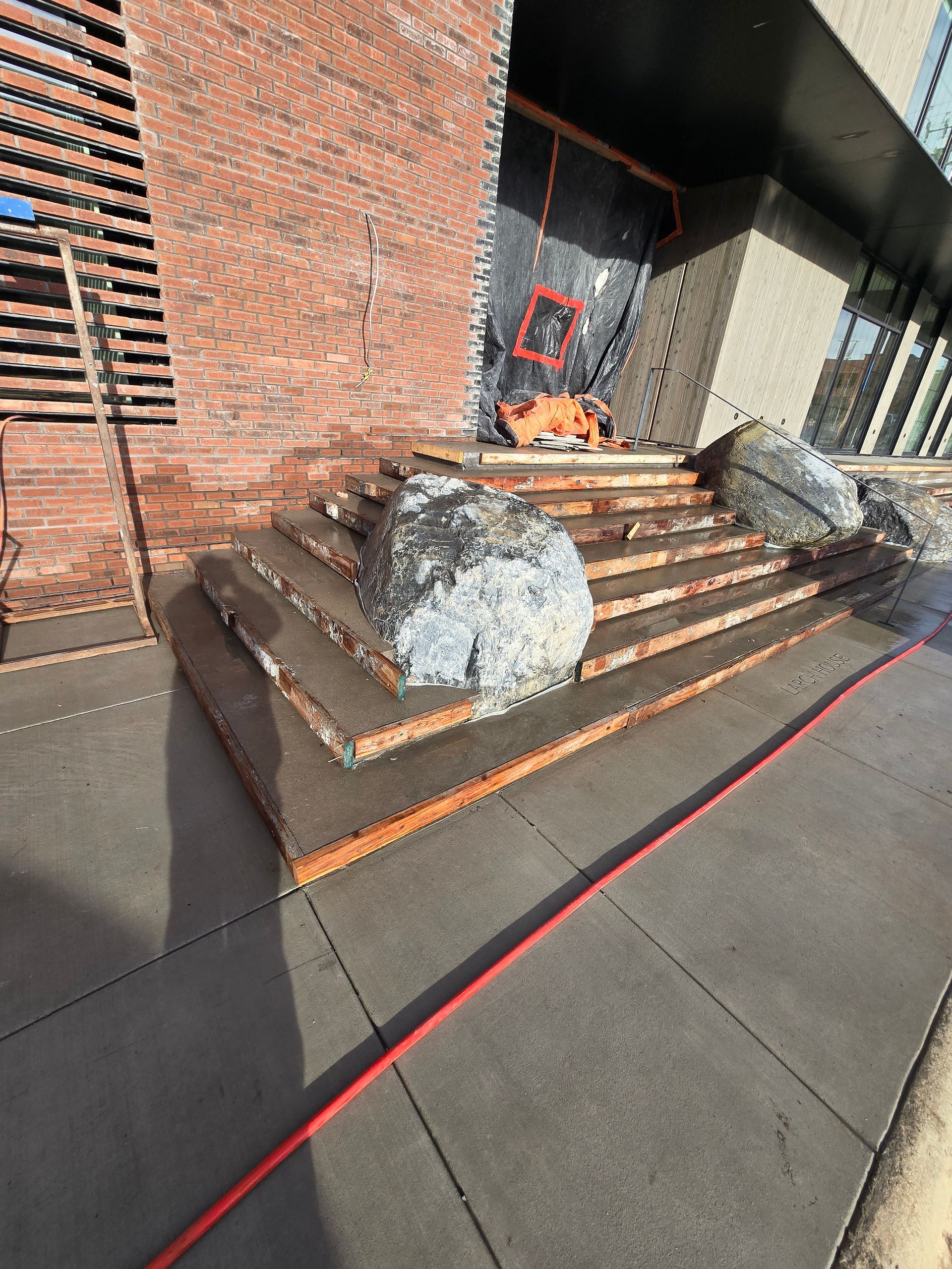 A construction site with concrete stairs flanked by two large boulders leading to a brick building entrance.