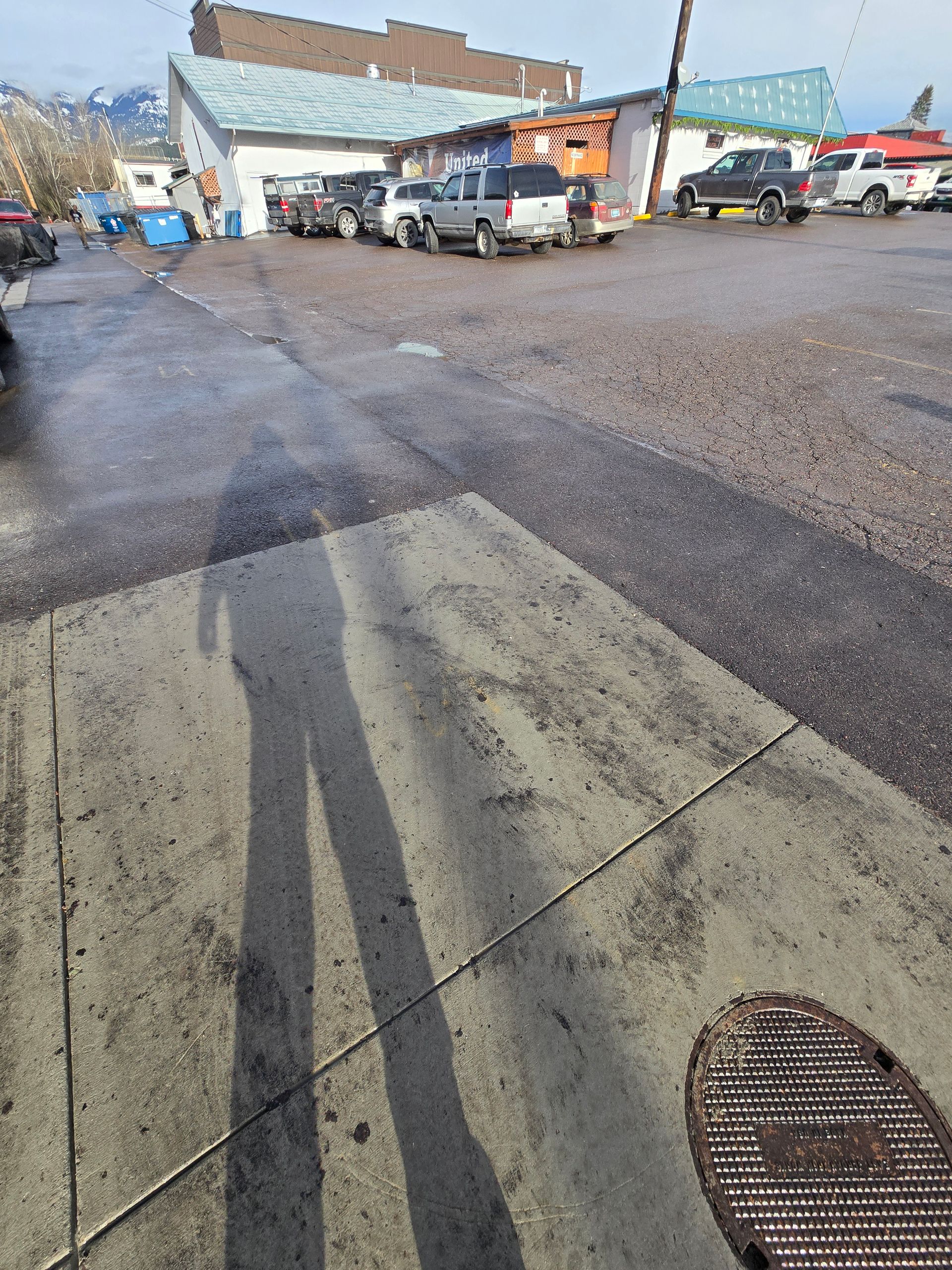 A long shadow of a person stands on a concrete sidewalk in front of a gravel parking lot with several parked vehicles.