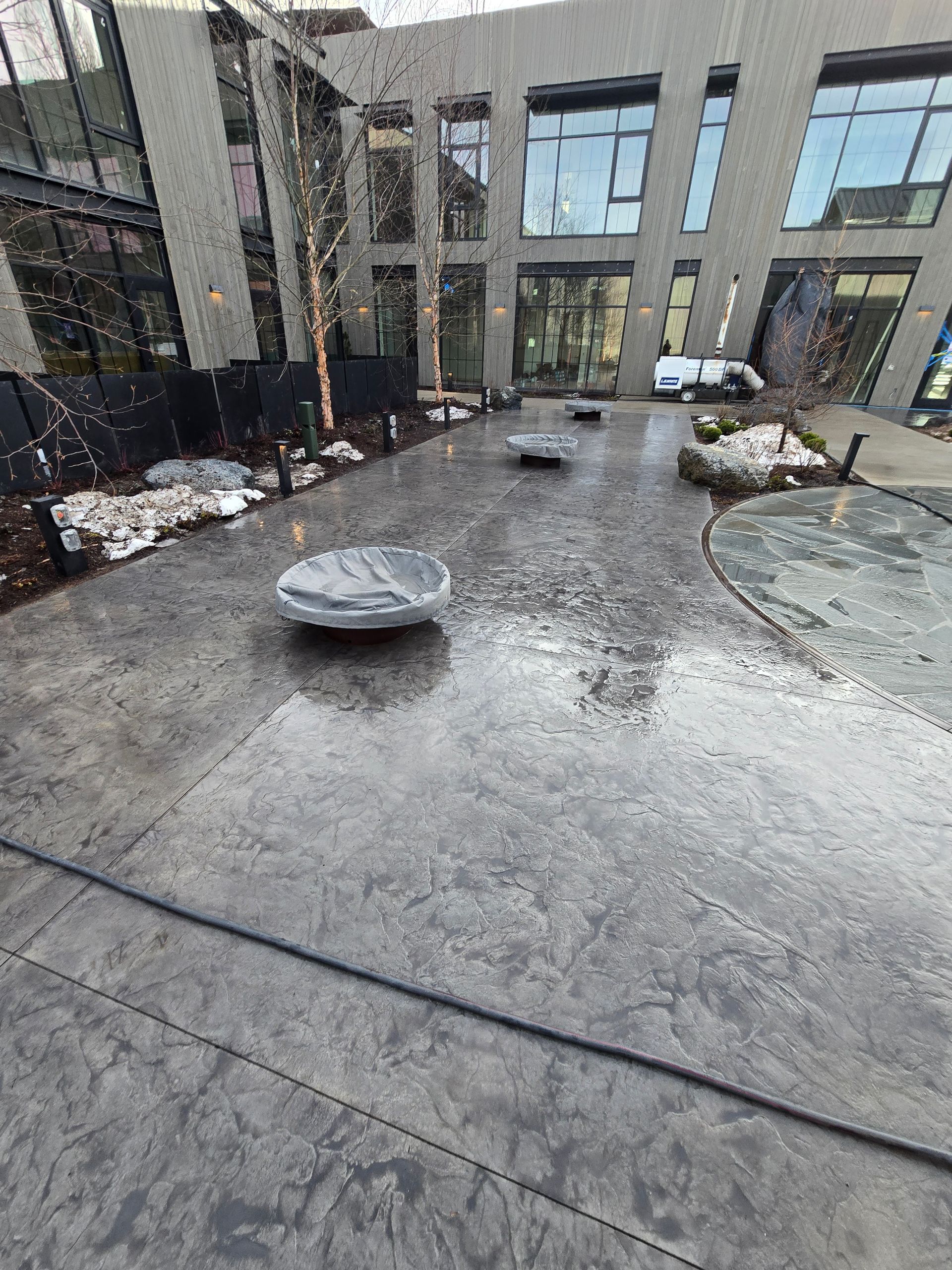 A grey stamped concrete patio outside a modern building, featuring low circular benches and decorative landscaping.