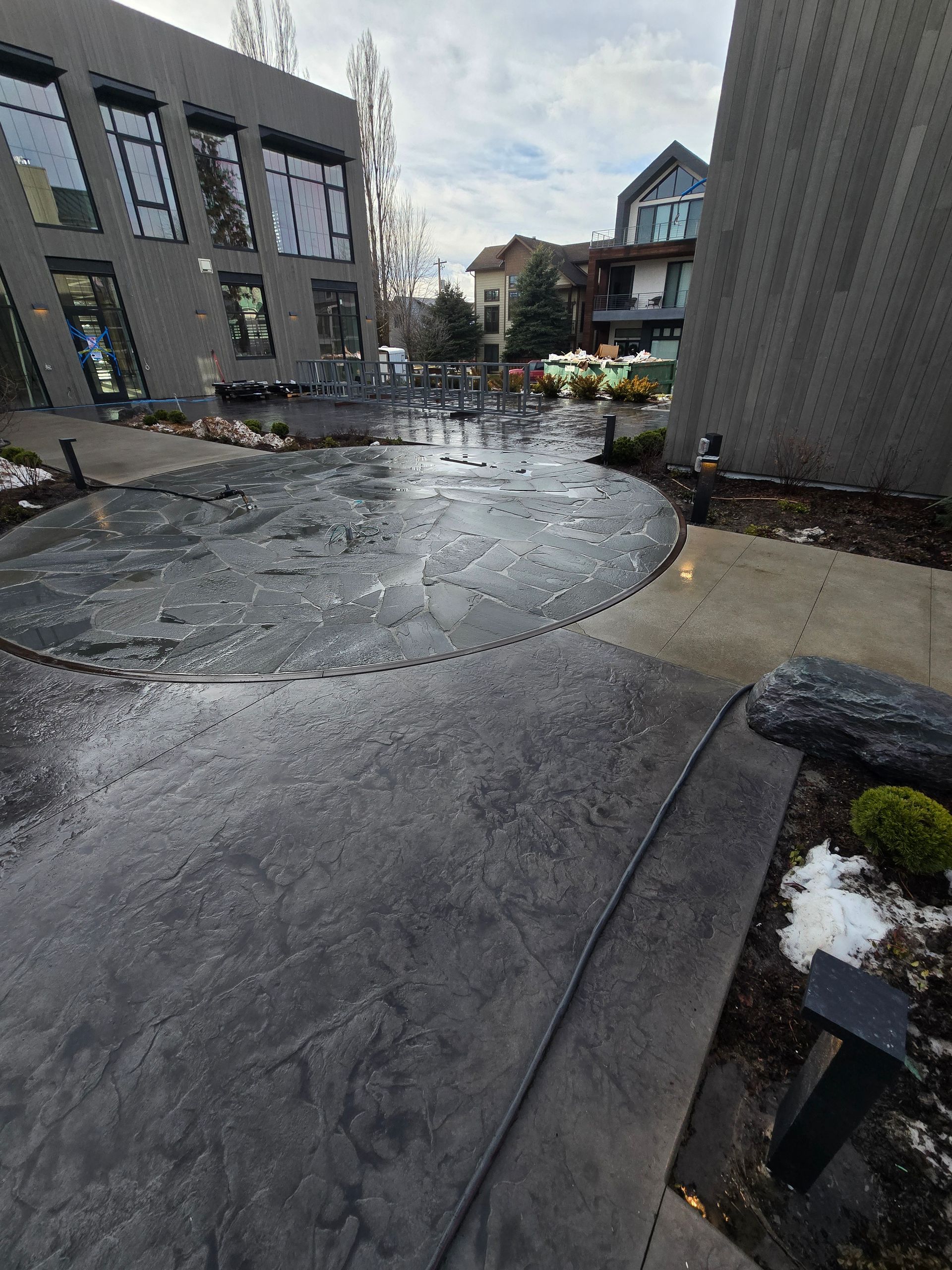 A wet paved courtyard between two modern buildings, featuring a circular textured patio area and sparse landscaping.