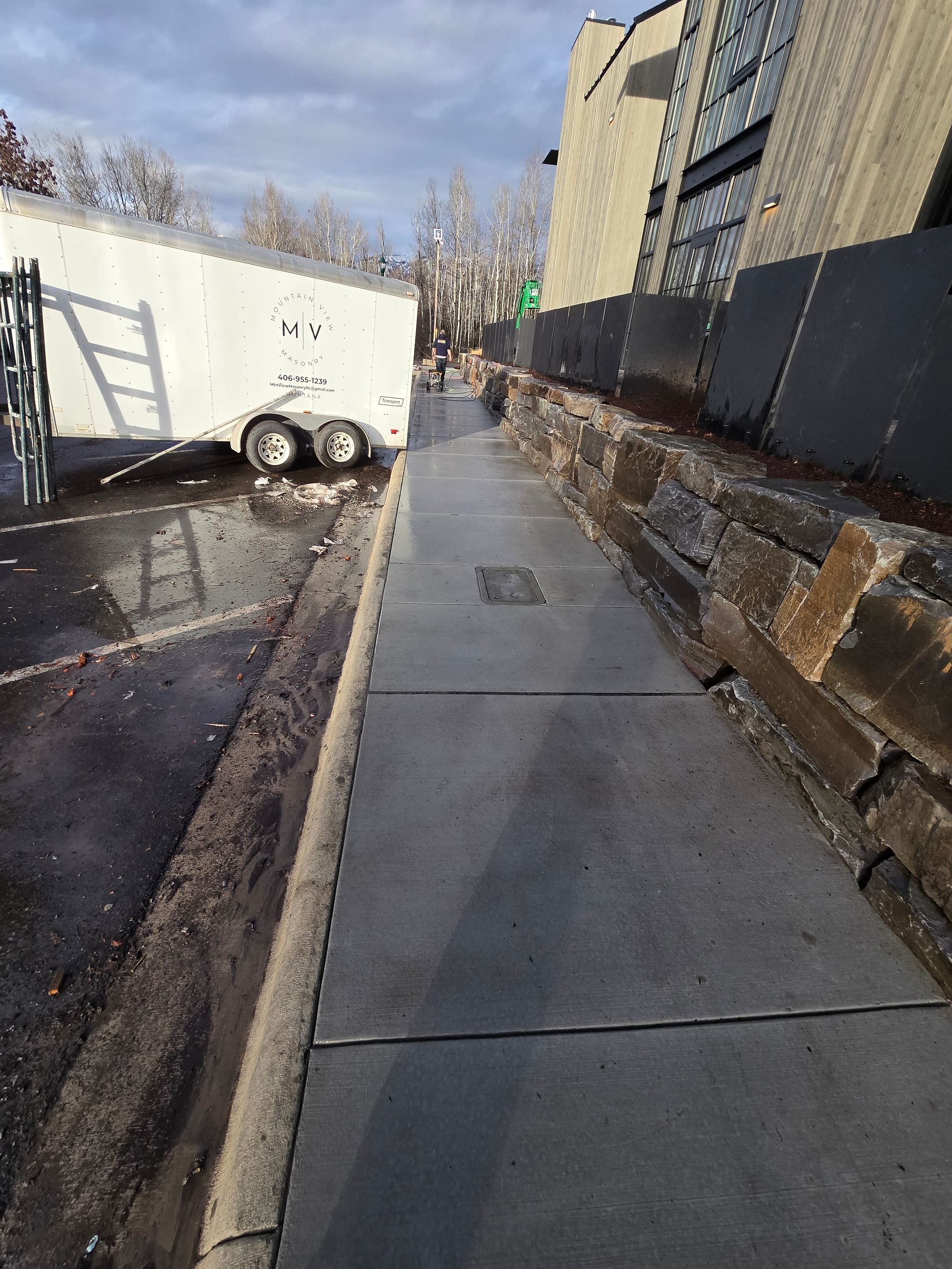 A sidewalk runs beside a stone retaining wall and a white trailer parked on asphalt under a cloudy sky.