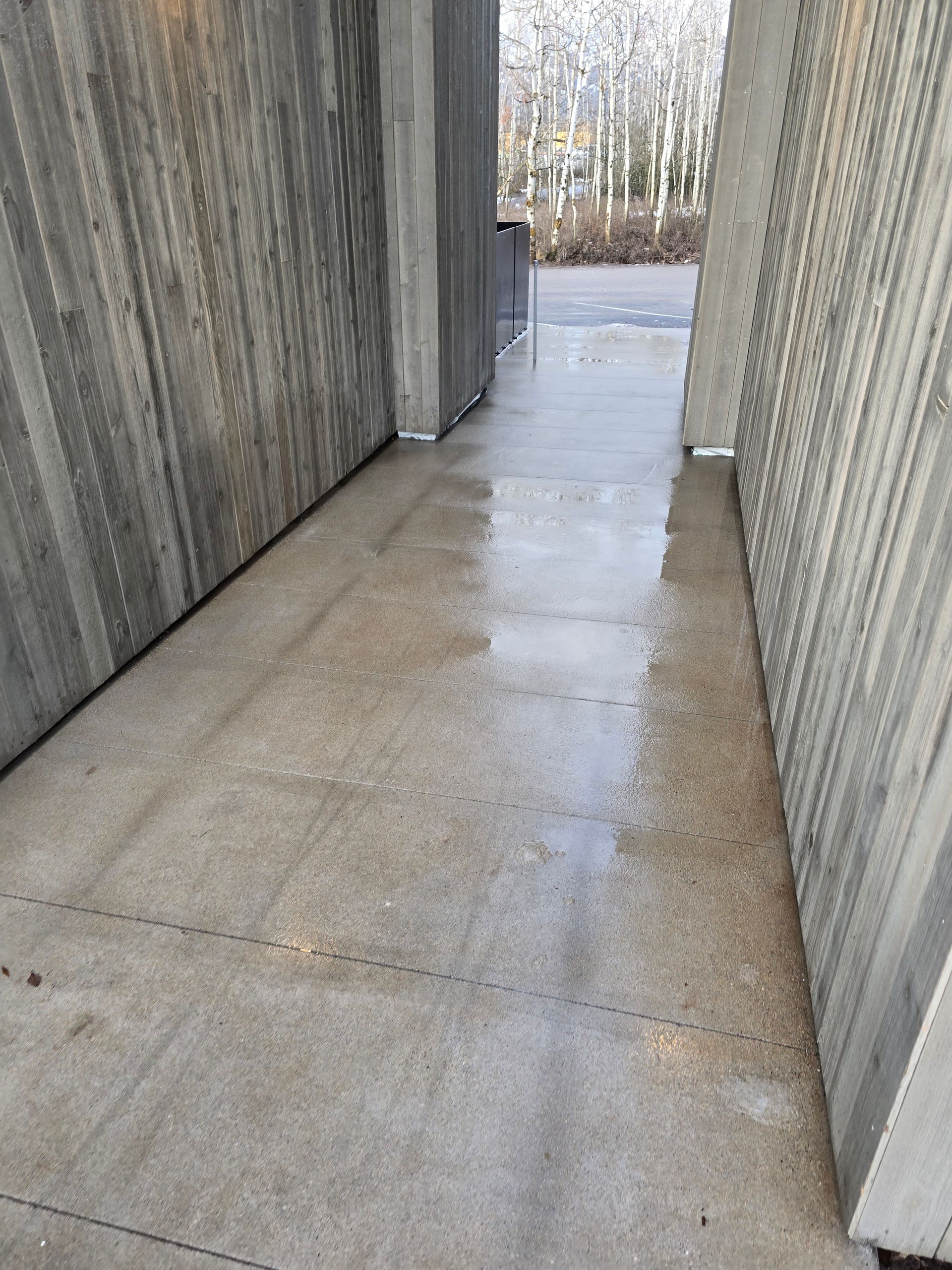 A narrow concrete path between two textured gray walls, with wet ground reflecting light leading toward an outdoor area.