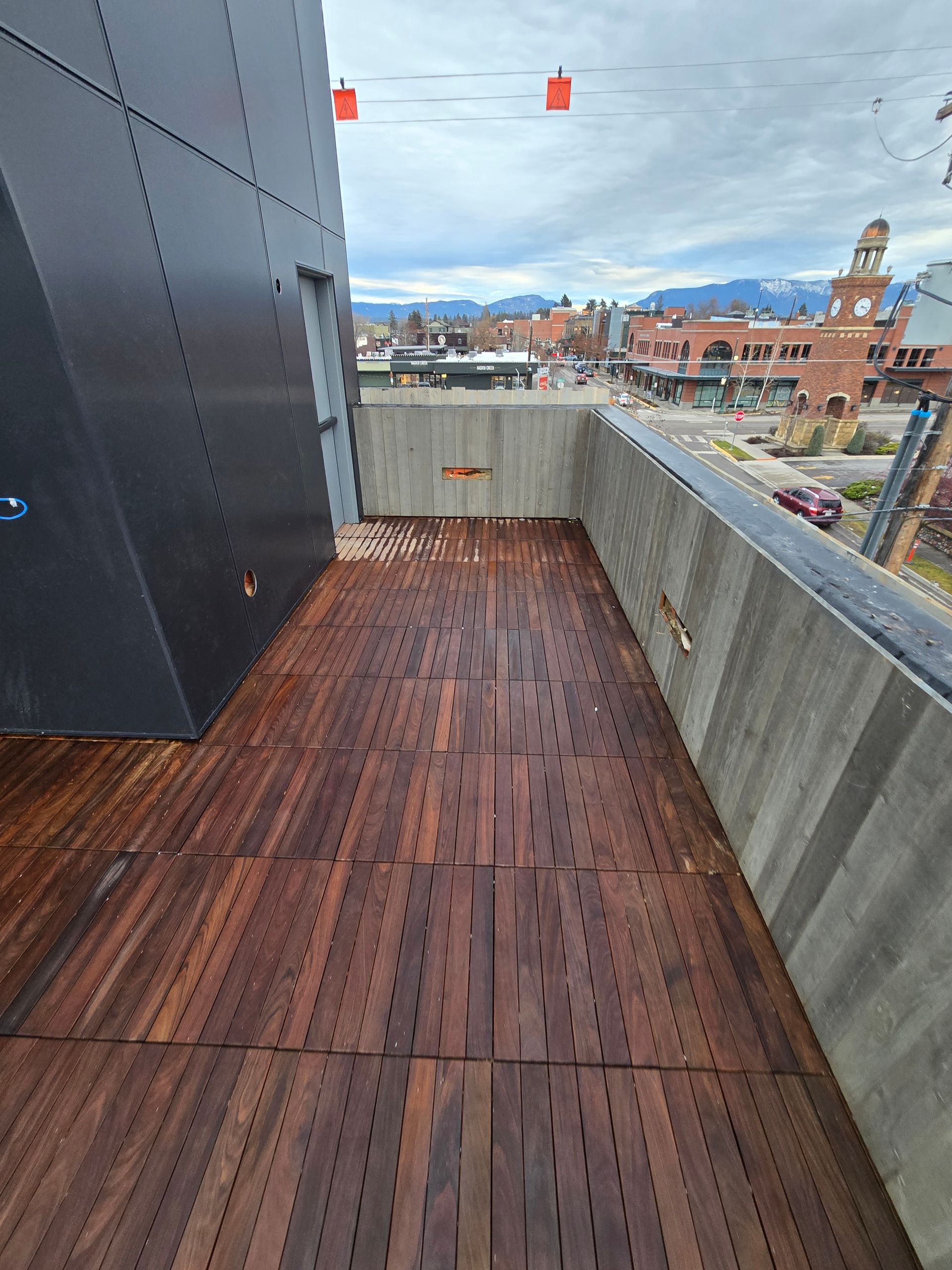 An outdoor balcony with dark brown wood-tiled flooring and concrete walls, overlooking a small town under a cloudy sky.