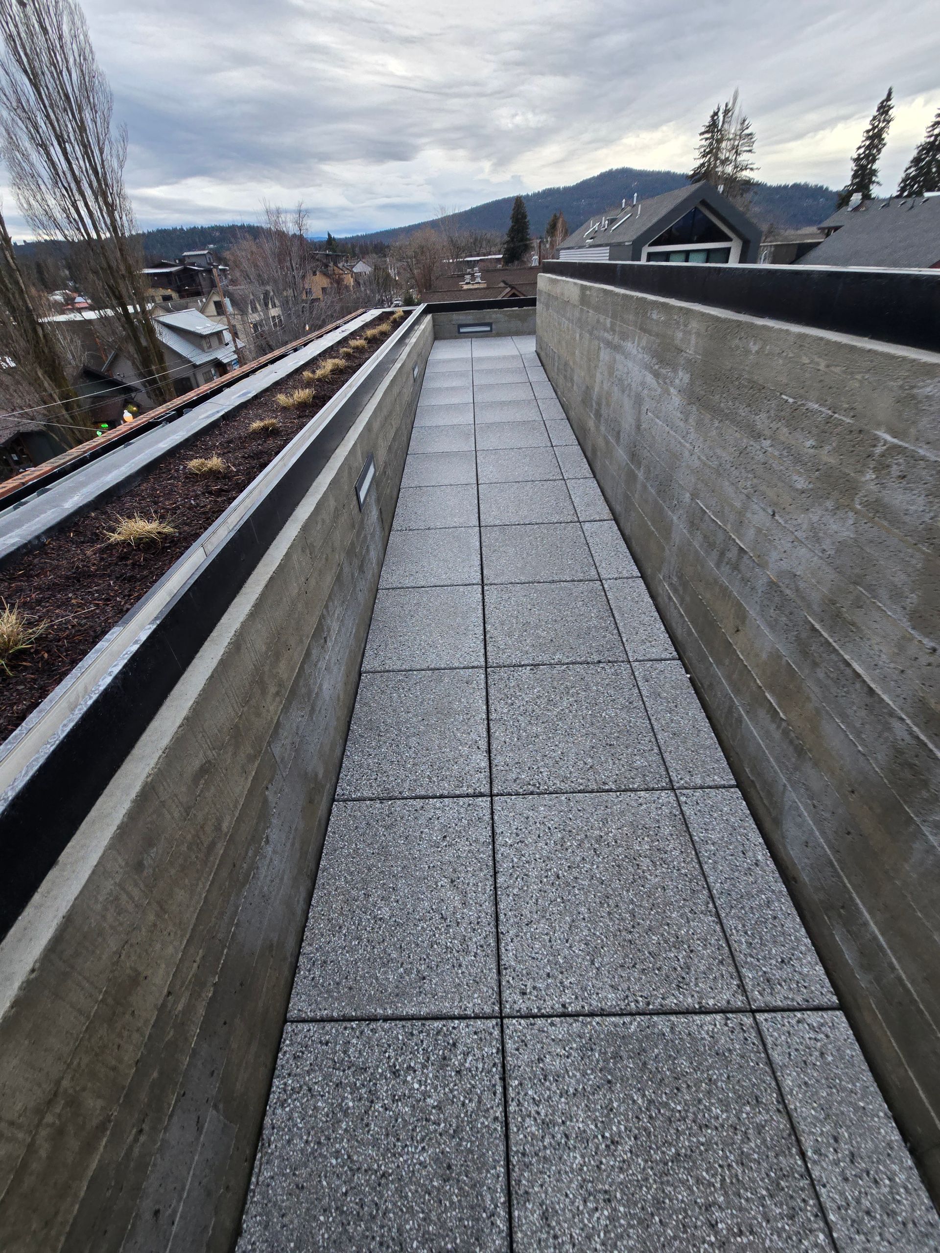 A narrow walkway with gray concrete pavers between a raised planter bed and a concrete wall, outdoors with mountain views.