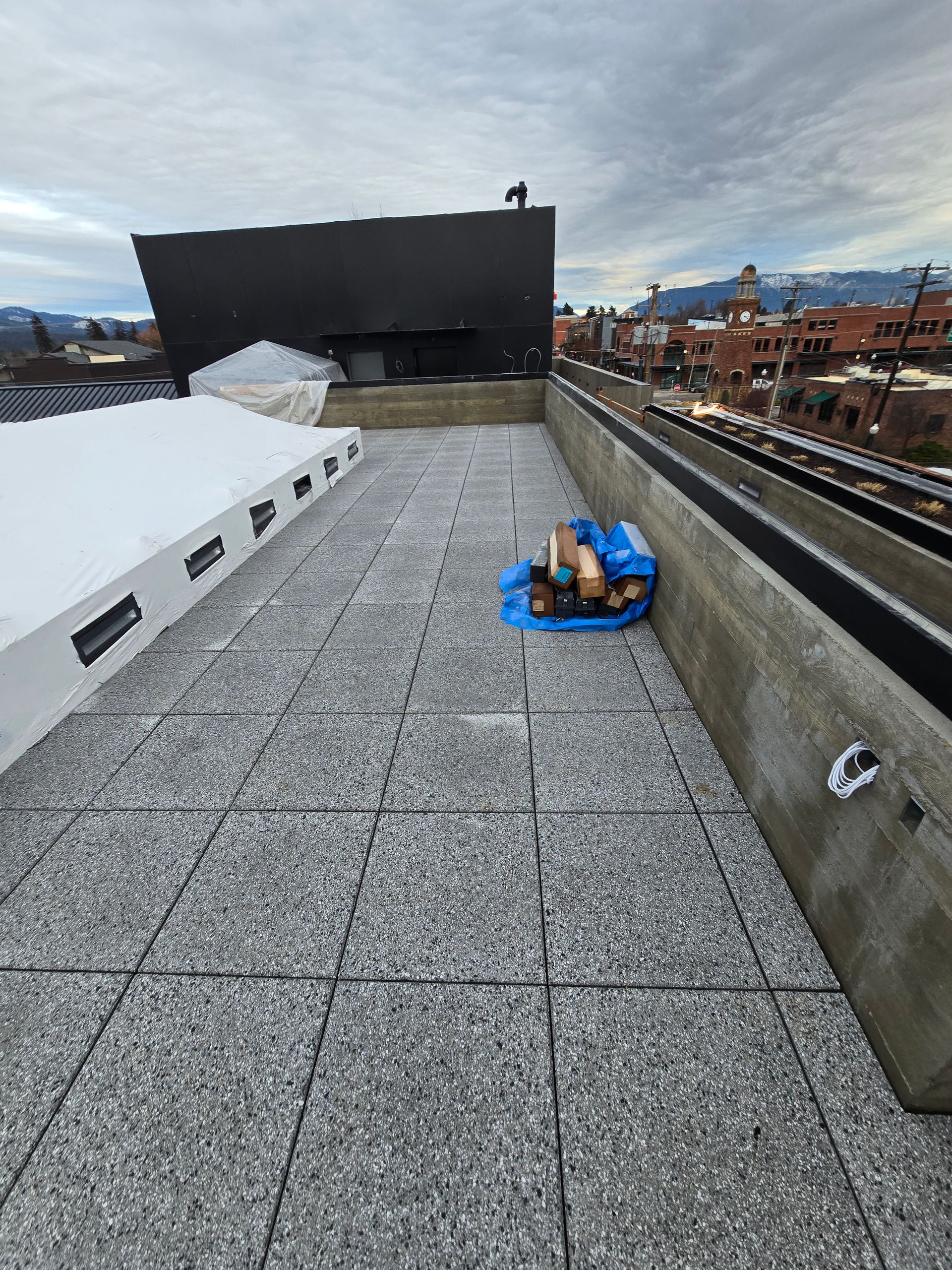 A rooftop patio with gray tiled flooring, a pile of wood on a blue tarp, and a large black building against a cloudy sky.