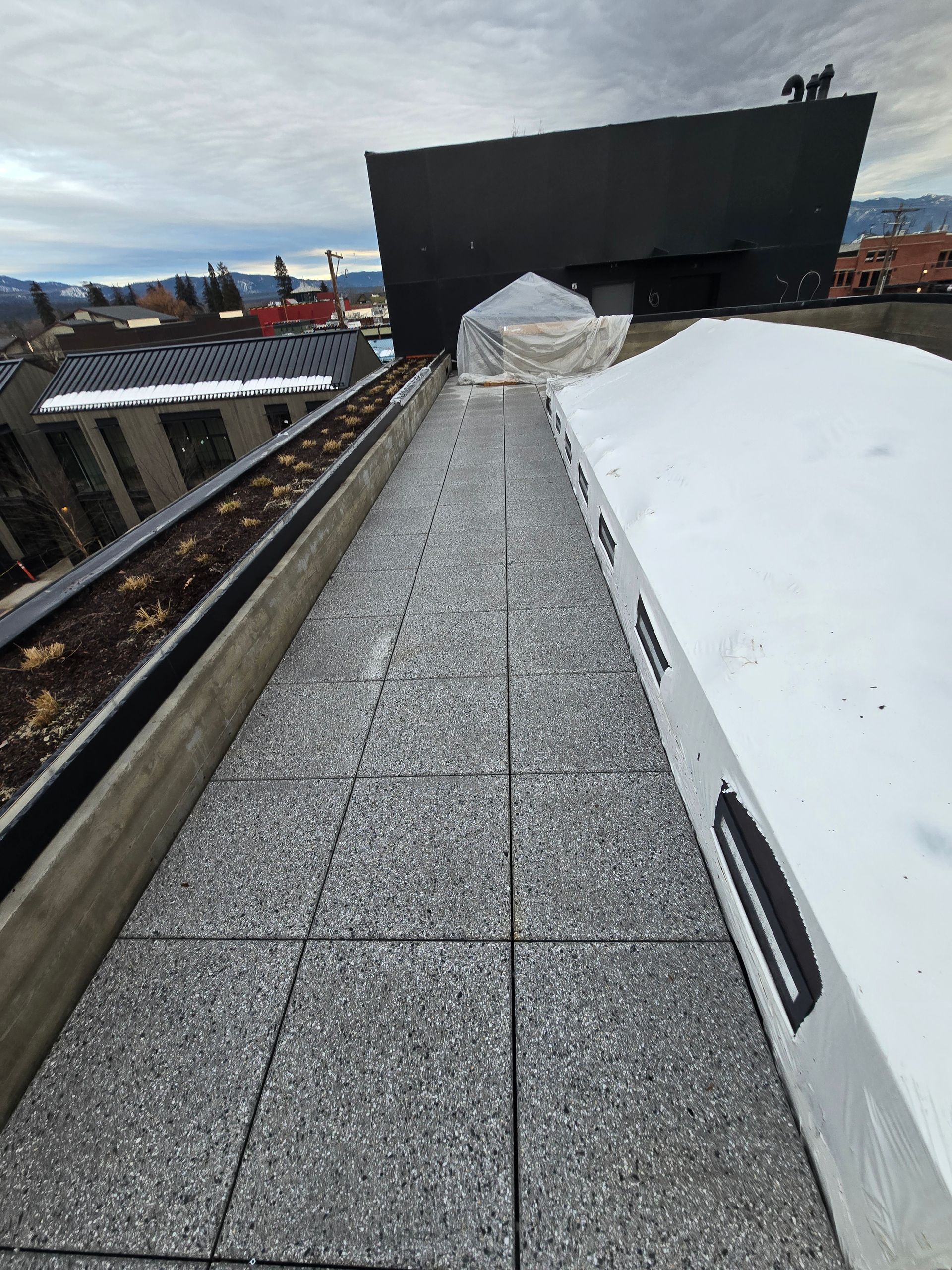 A rooftop walkway with textured grey tiles, bordered by a snow-covered area and a planter box against a dark building wall.