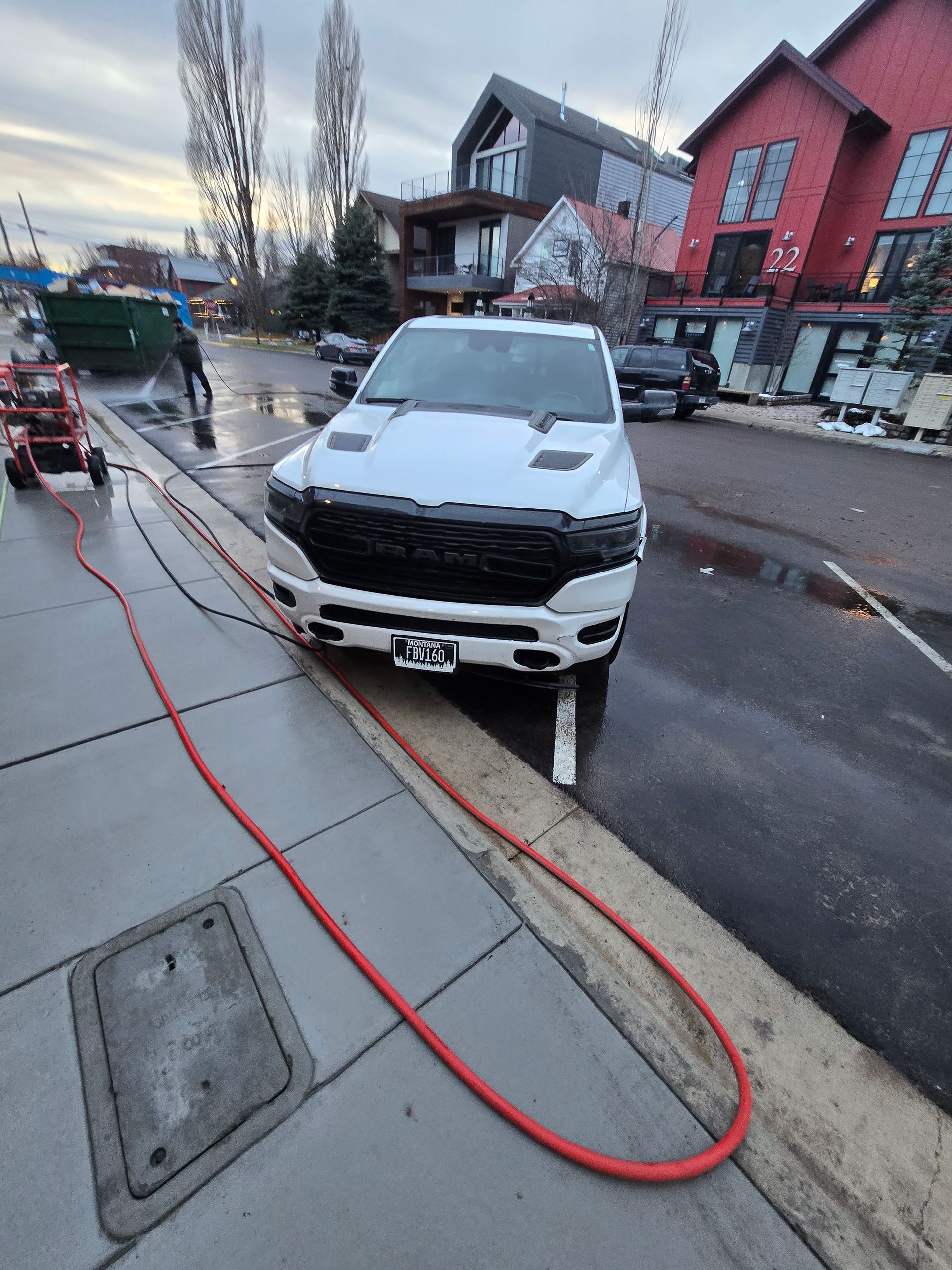 A white pickup truck parked on a wet street beside a sidewalk with a long red hose extending from a nearby piece of gear.