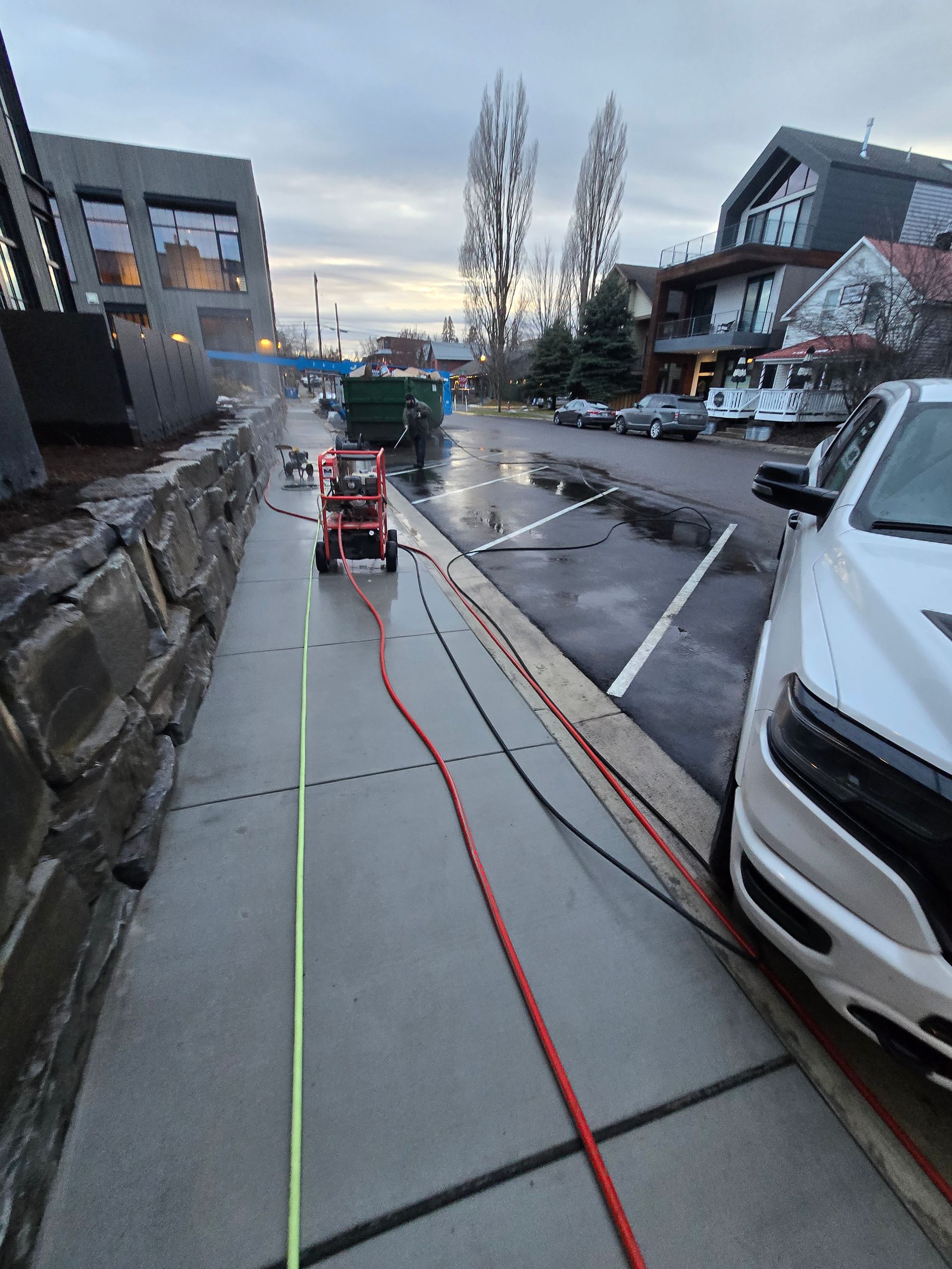 A red pressure washer sits on a sidewalk next to a white truck, with hoses running along the path in a residential area.