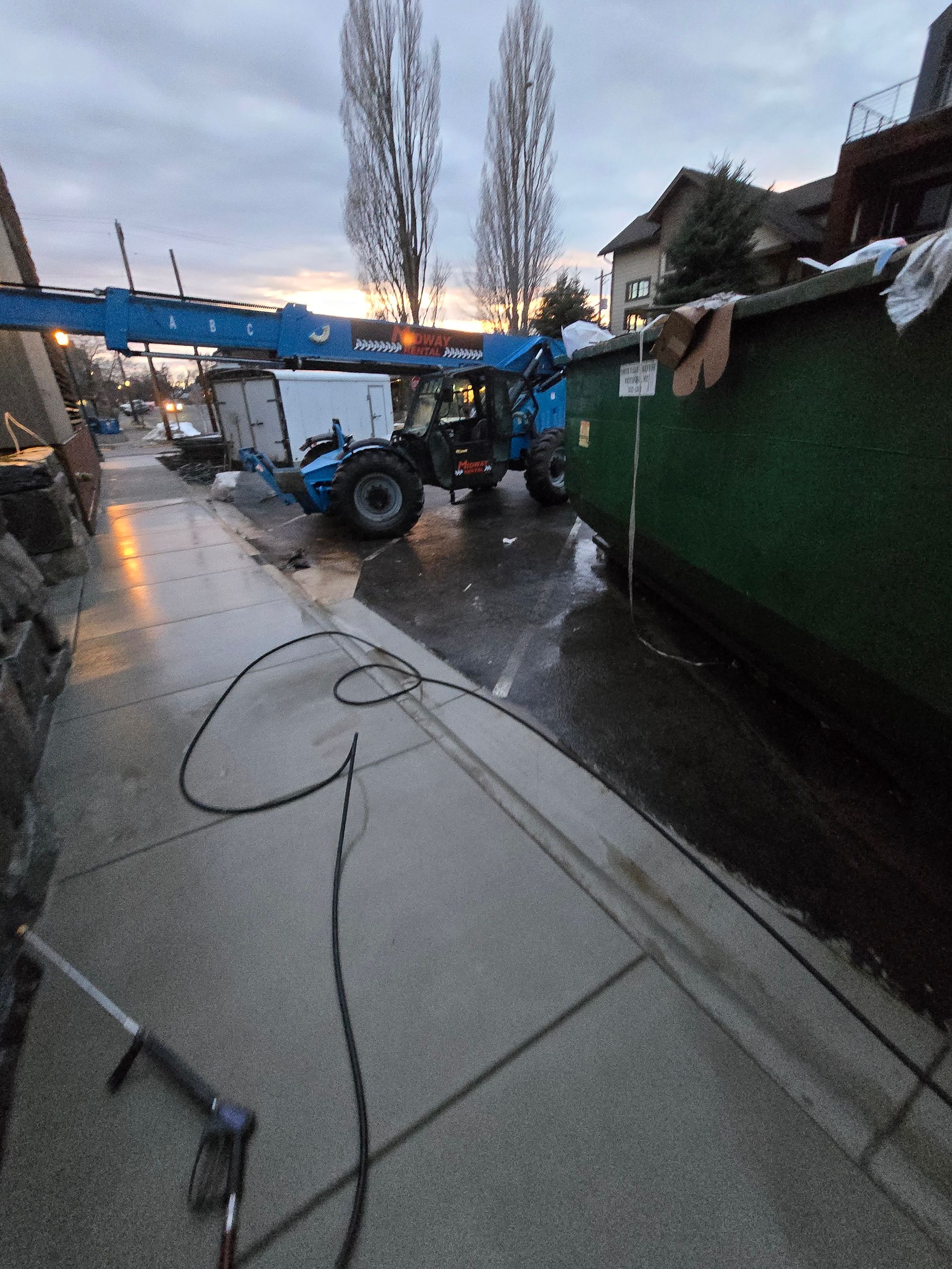 A blue telescopic handler parked on a street near a large green dumpster at dusk, with a spray nozzle on the sidewalk.