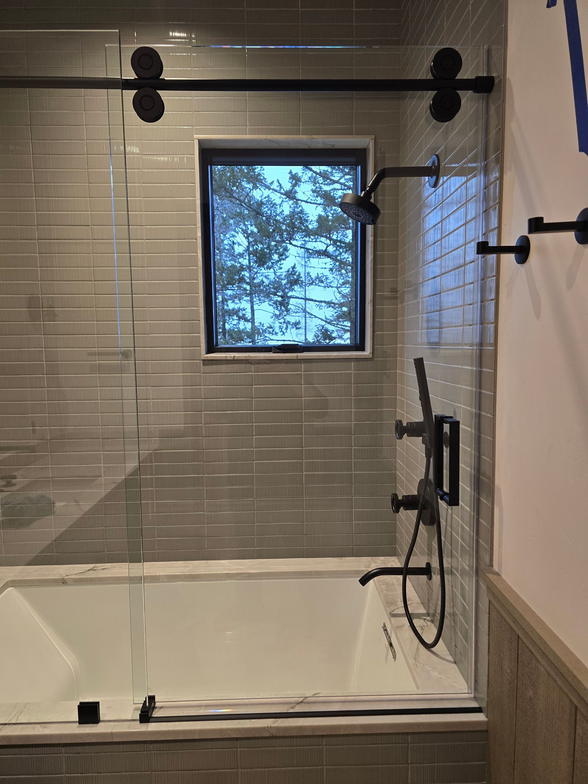A white bathtub with matte black fixtures and a glass sliding door against a gray tiled wall with a small window.