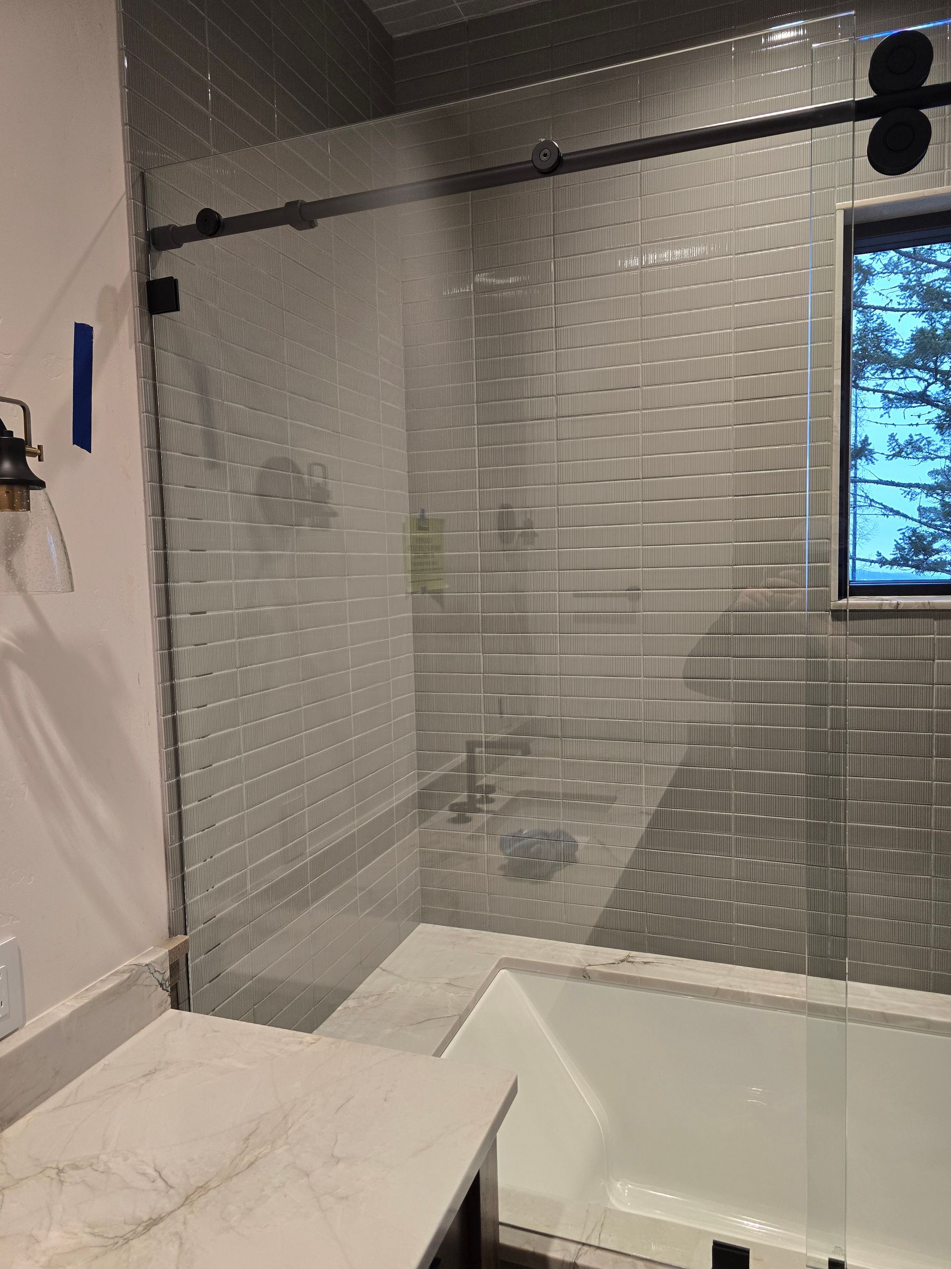 A sliding glass shower door installed in a bathroom featuring gray tiled walls and a white bathtub.