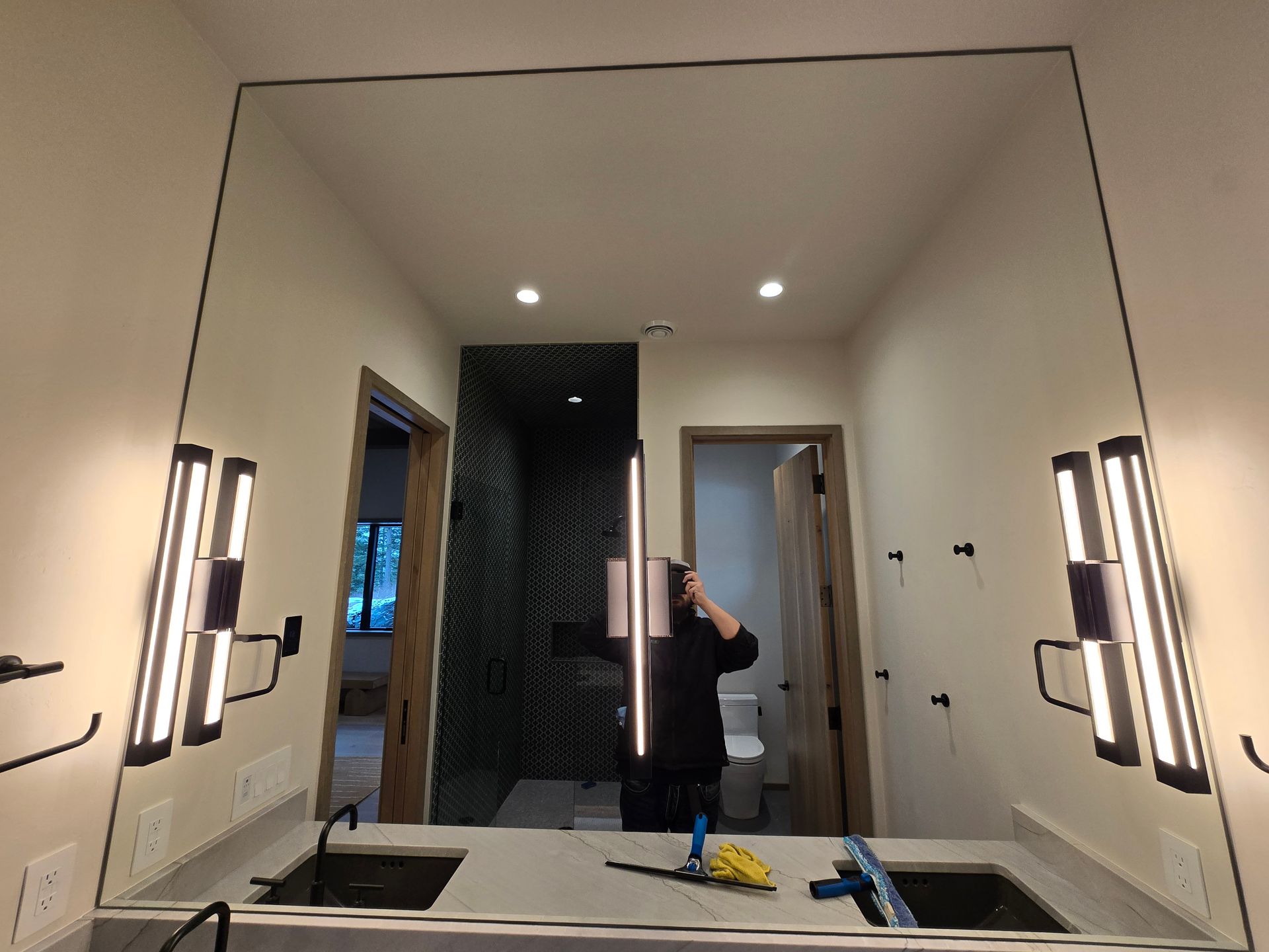 A large rectangular bathroom mirror reflects a person holding a phone, two modern vertical wall lights, and a doorway.